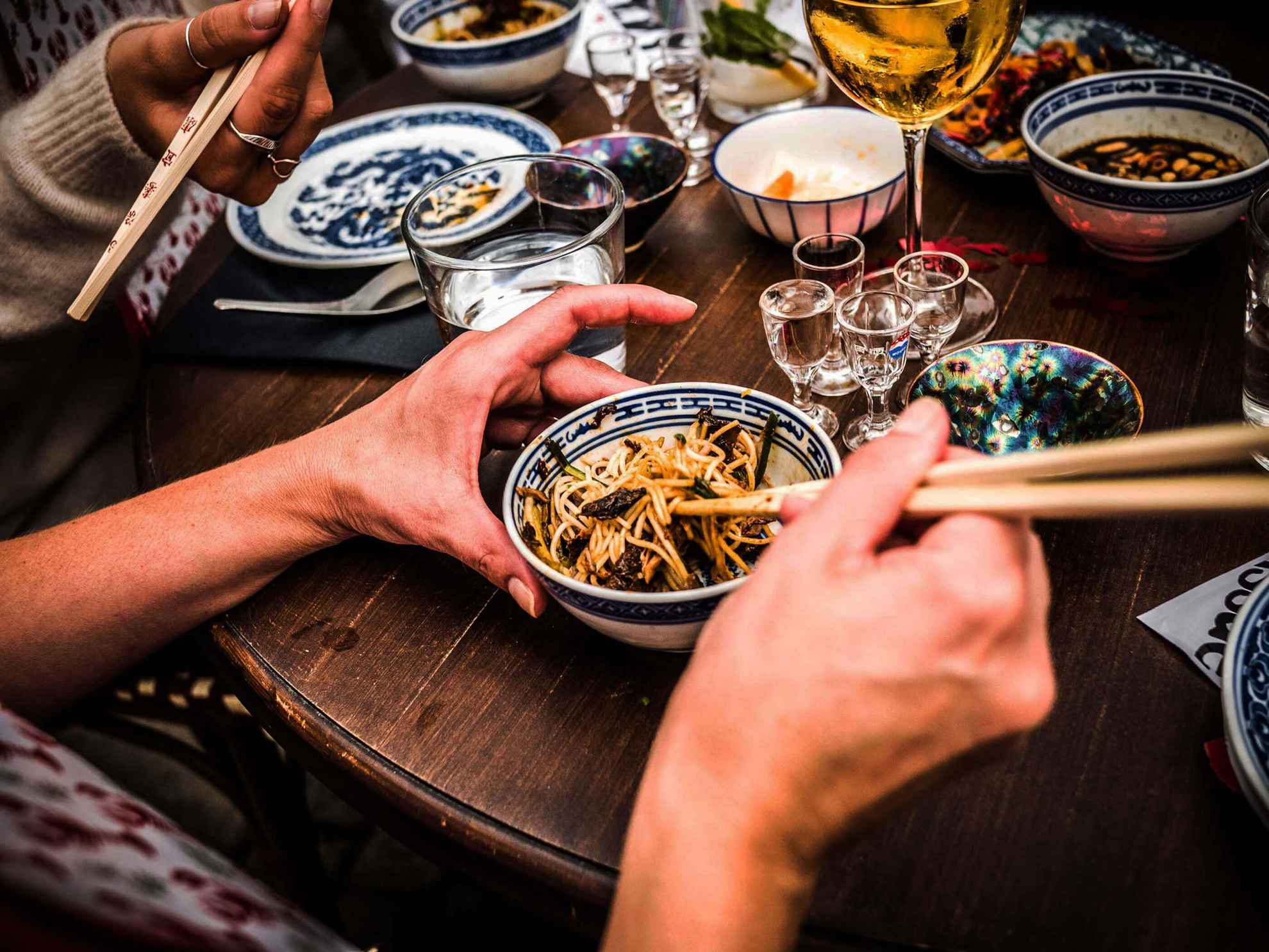 Ein Tisch gedeckt mit Gerichten. Menschen essen mit Essstäbchen aus kleinen verzierten Schalen.
