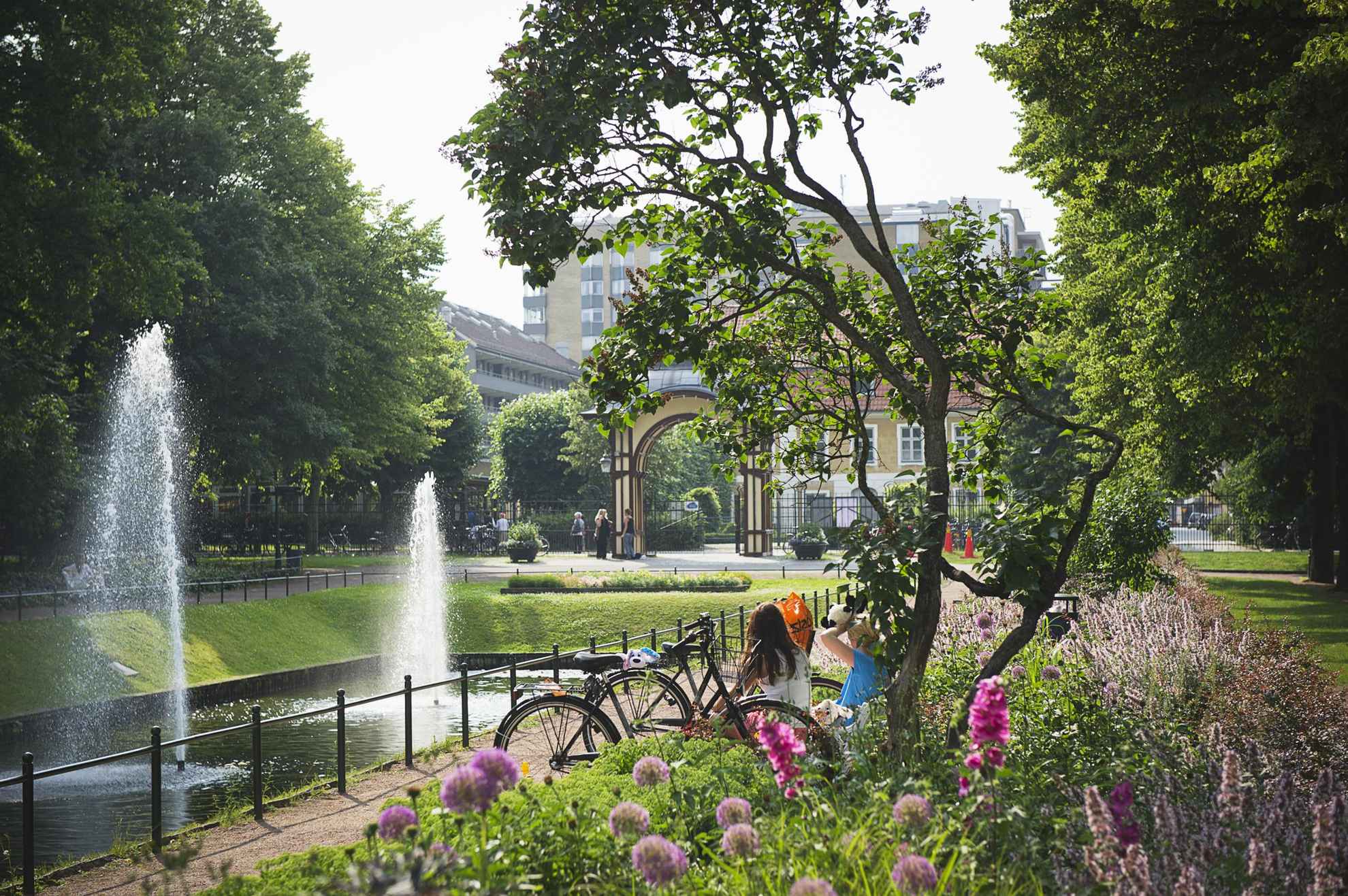 Blumen und Grün in einem Stadtpark. Man sieht den Rücken von zwei Frauen, die neben einem Becken mit zwei Springbrunnen sitzen. Im Hintergrund sieht man den Eingang und einige Gebäude außerhalb des Parks.
