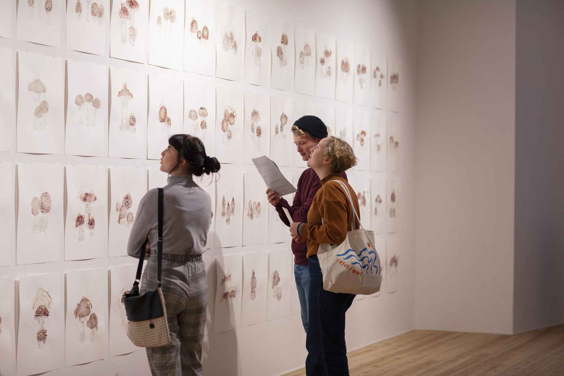 Ein Mann und zwei Frauen betrachten mehrere Gemälde von Pilzen an einer Wand in einem Museum.