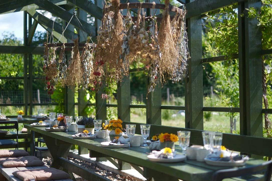 Ein grüner langer Holztisch ist mit Gläsern, Tellern und gelben Blumen gedeckt. Der Tisch steht im Freien in einem Holzgebäude.