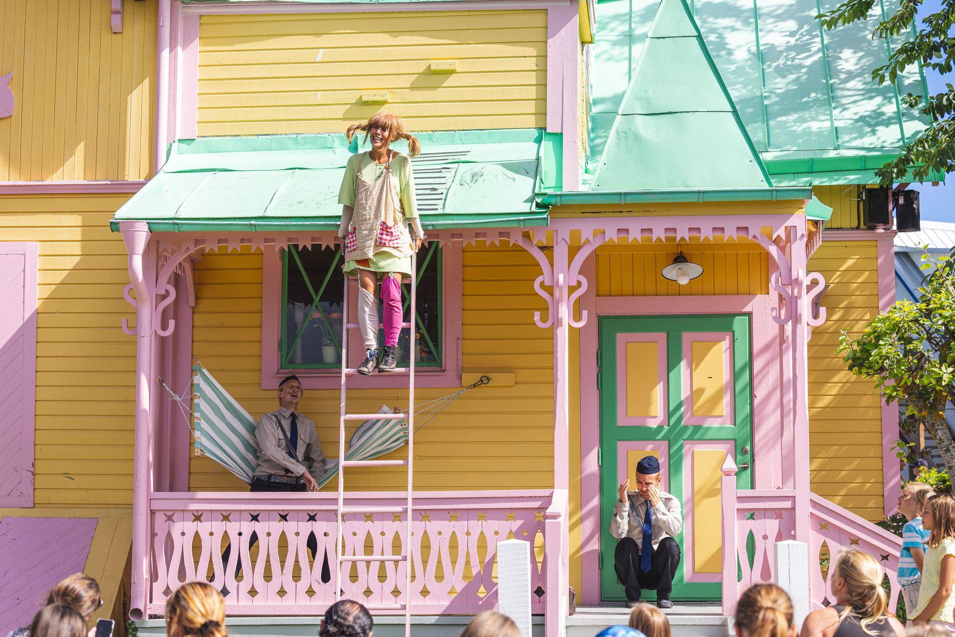 Das gelbe Haus von Pippi Langstrumpf, mit Publikum vor dem Haus, das eine Theateraufführung mit Pippi sieht, die auf einer Leiter steht.