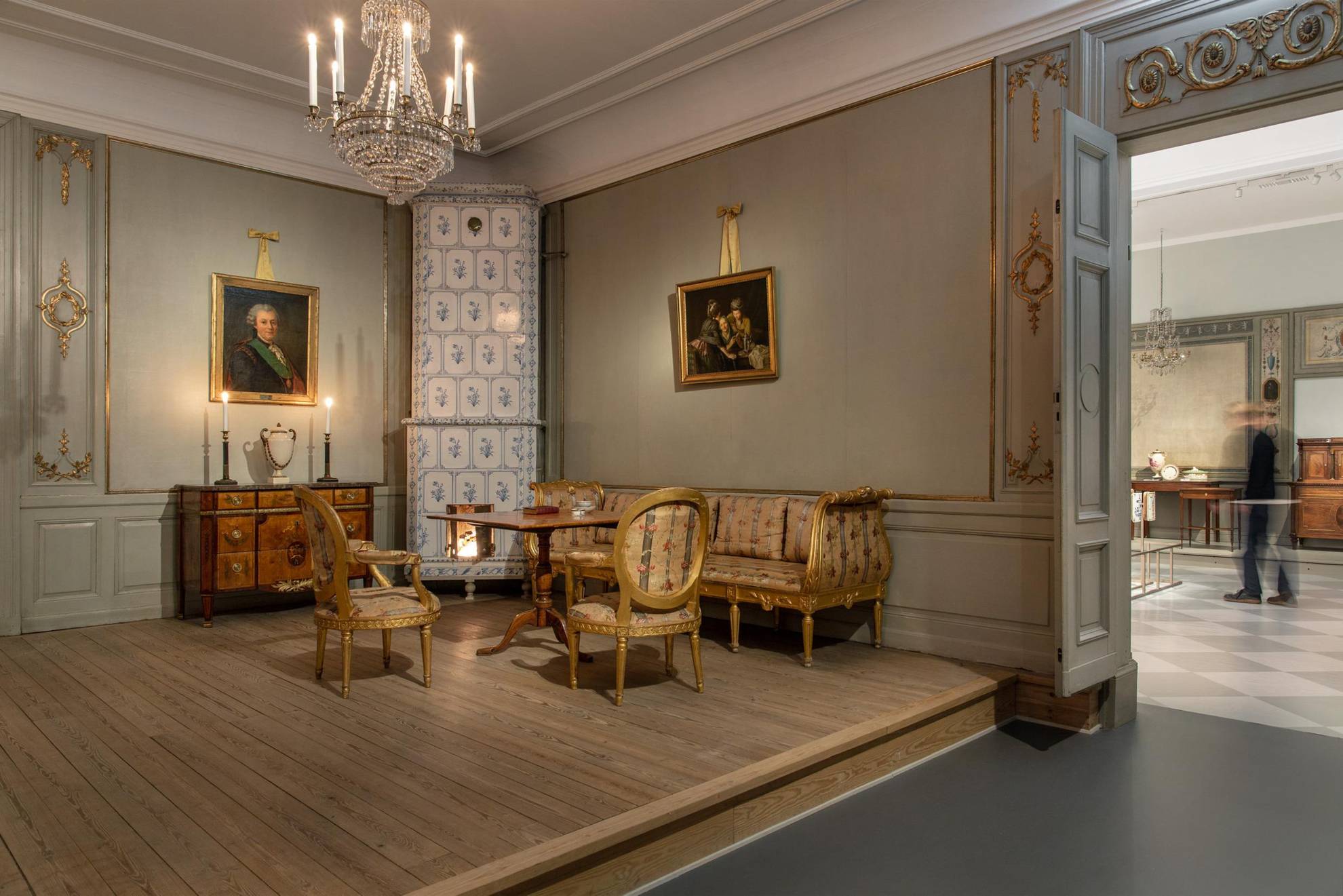 Möbel aus dem 18. Jahrhundert in einer Ausstellung. Ein Kachelofen, ein Sofa und zwei Stühle, ein Tisch und eine Kommode, ein Kristallkronleuchter, der von der Decke hängt, und zwei Gemälde an der Wand.