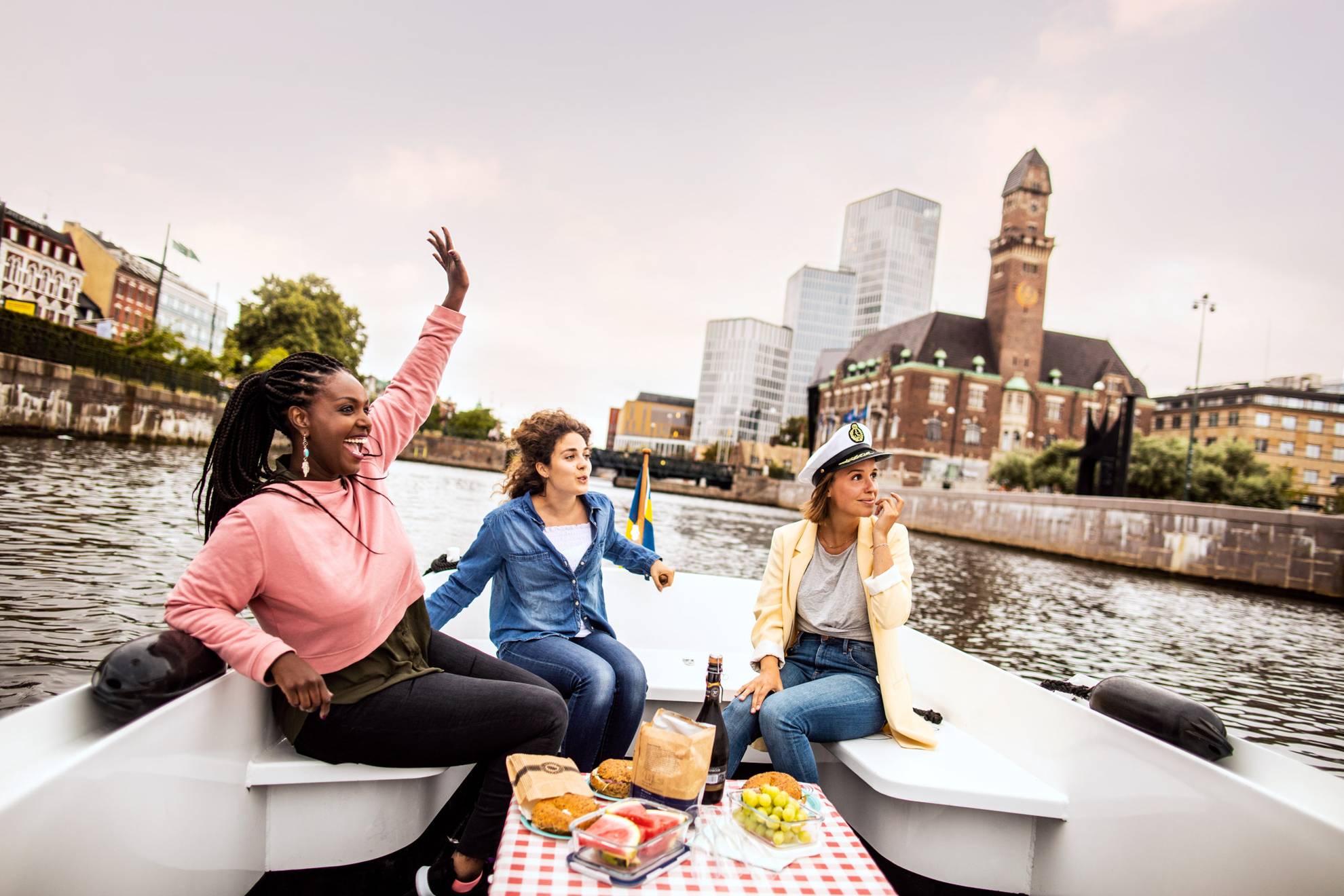 Drei junge, glückliche Frauen bei einem Ausflug in einem Boot auf dem Malmöer Kanal mit Malmö Live und dem Hauptbahnhof im Hintergrund.