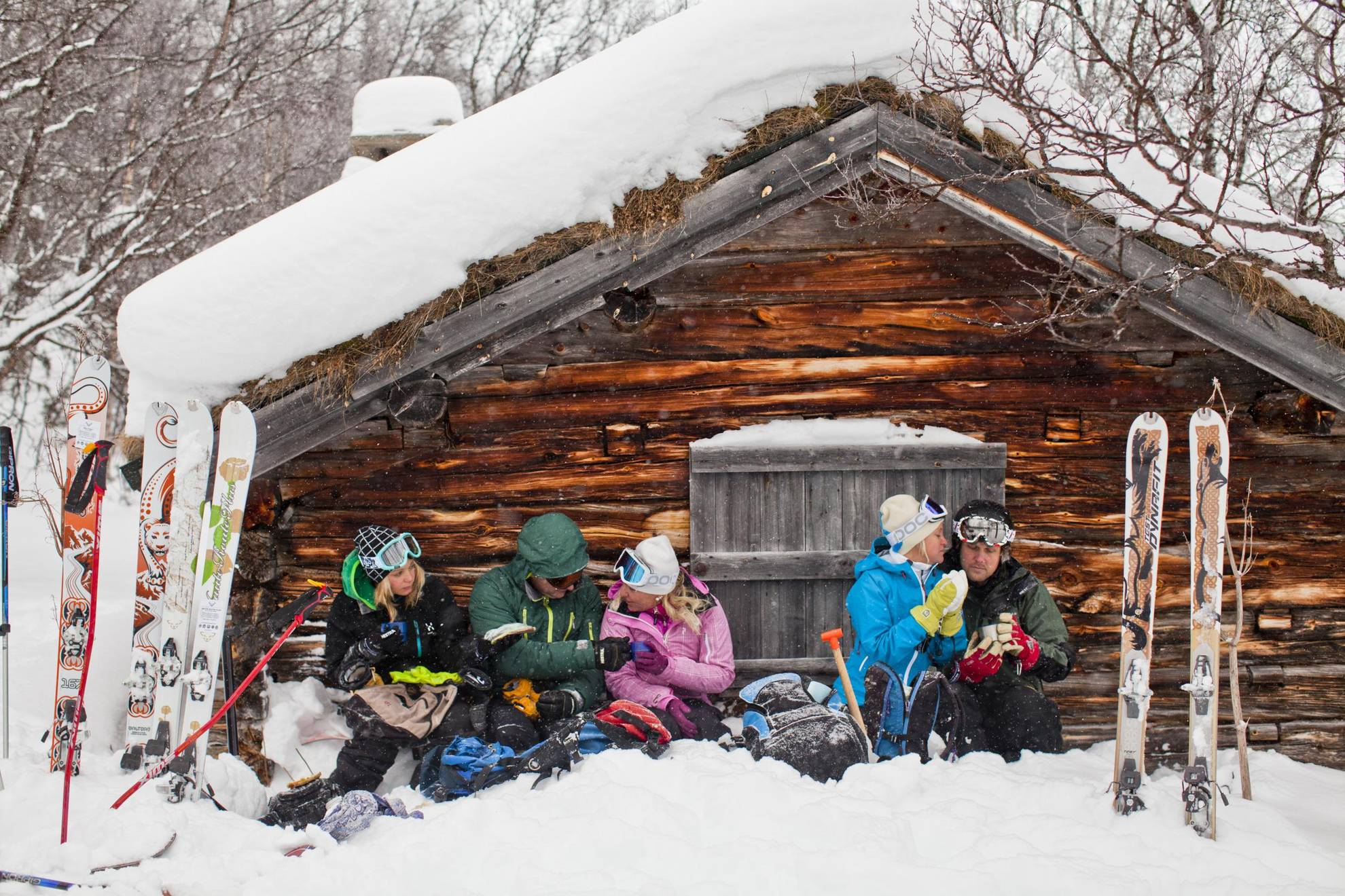 Fünf Leute sitzen im Schnee vor einem Holzhaus in den Bergen und machen eine Kaffeepause vom Skifahren.