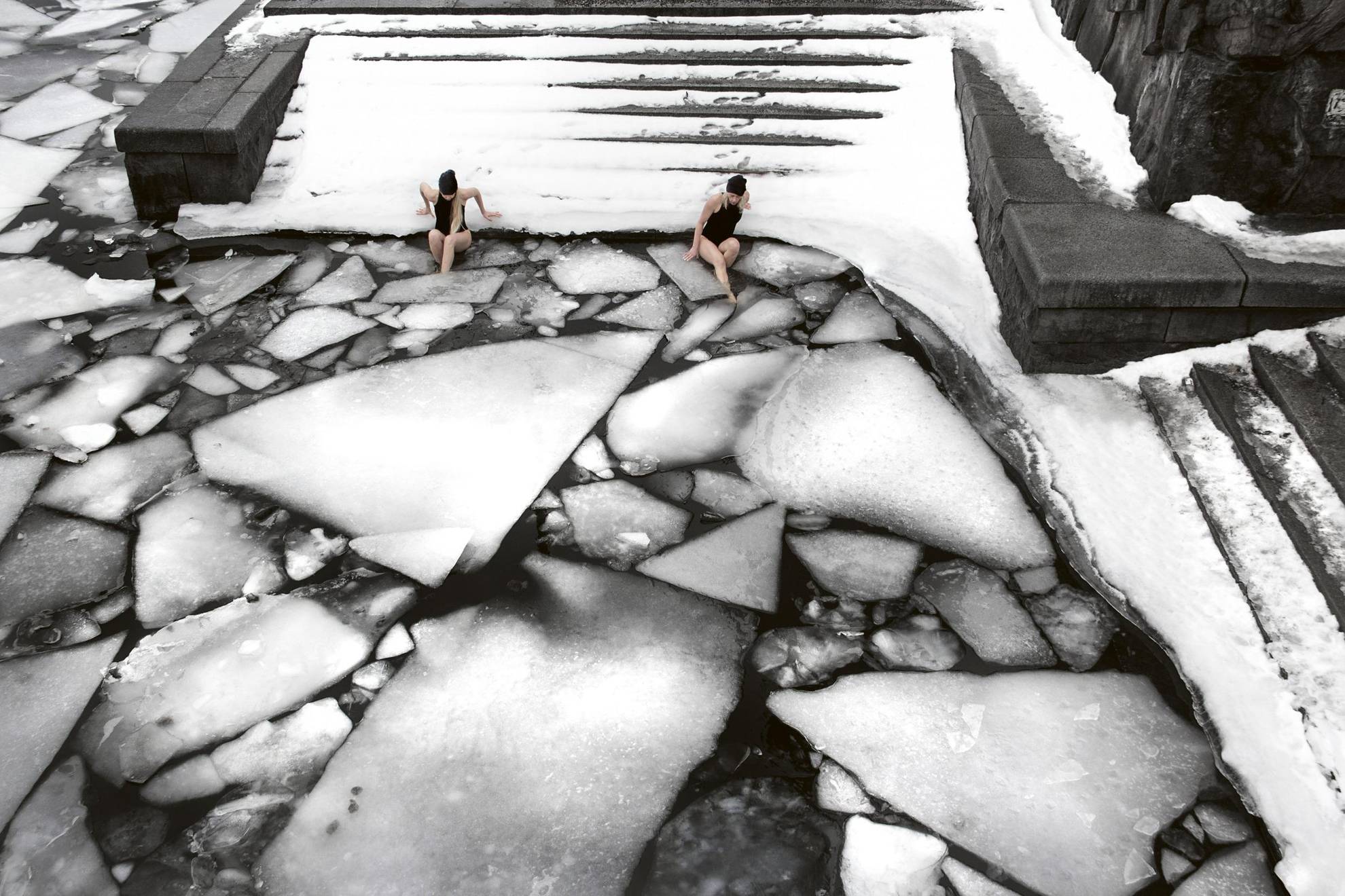 Zwei Frauen sind im Begriff, ein  Winterbad von einem Steg mit Treppe zum Ufer hinunter zu nehmen. Es gibt Eisschollen im Wasser.