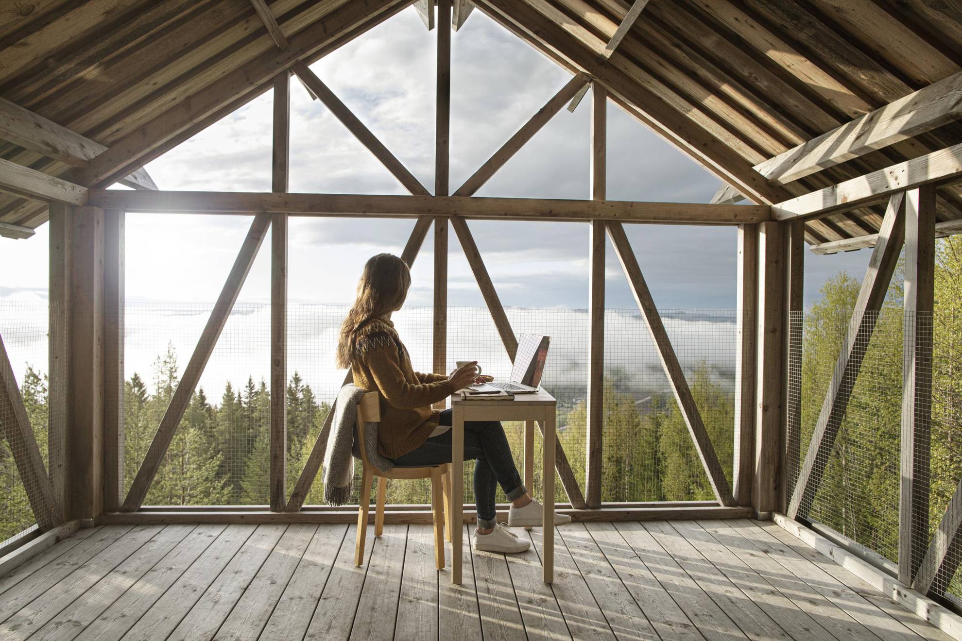 Eine Frau mit einer Tasse Kaffee sitzt an einem Schreibtisch in einem Raum mit Panoramafenstern mit Blick auf die spektakuläre Aussicht auf die umliegende Natur.