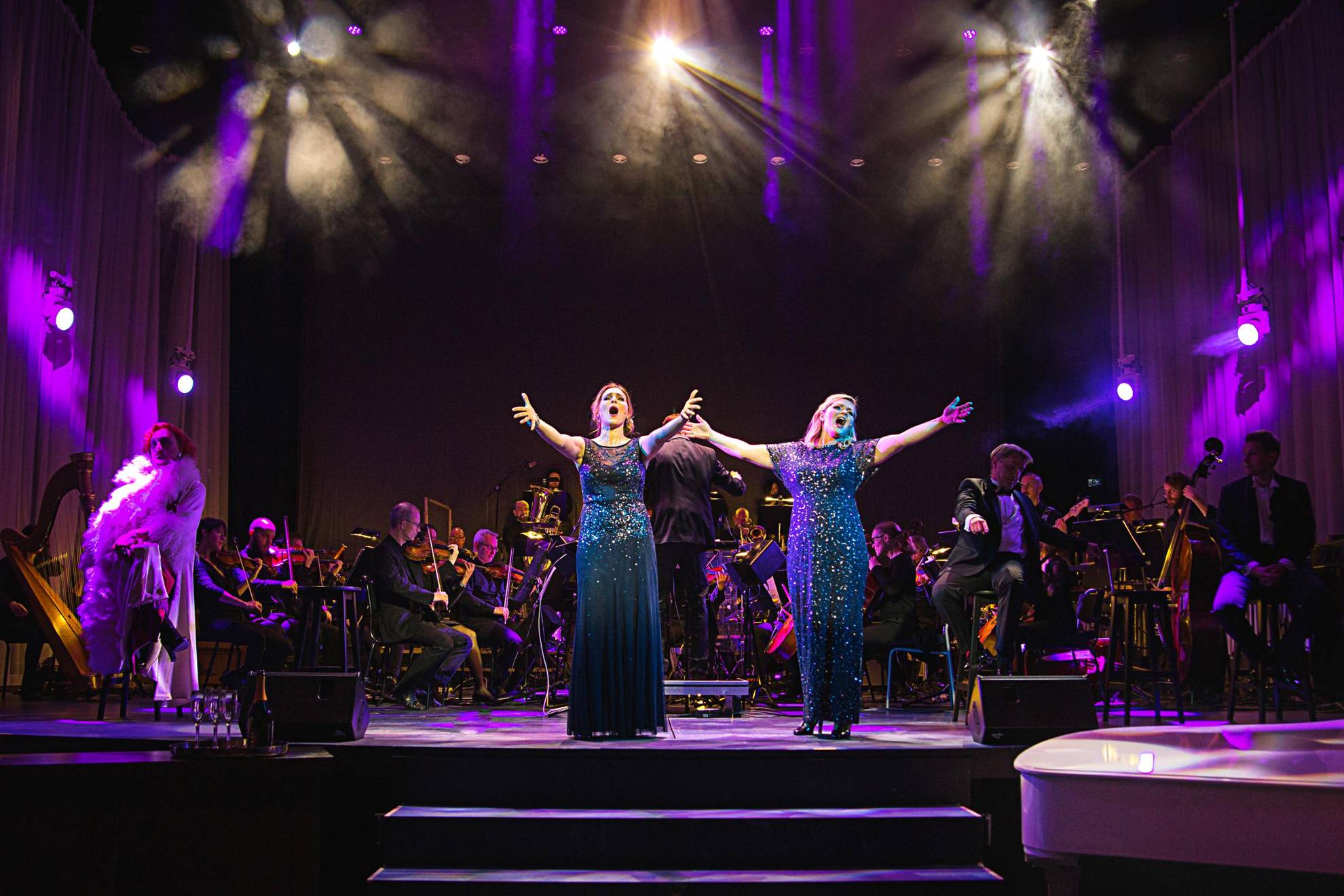 Zwei Frauen in blauen Glitzerkleidern singen während einer Aufführung auf einer Bühne. Dahinter sitzt ein Orchester.