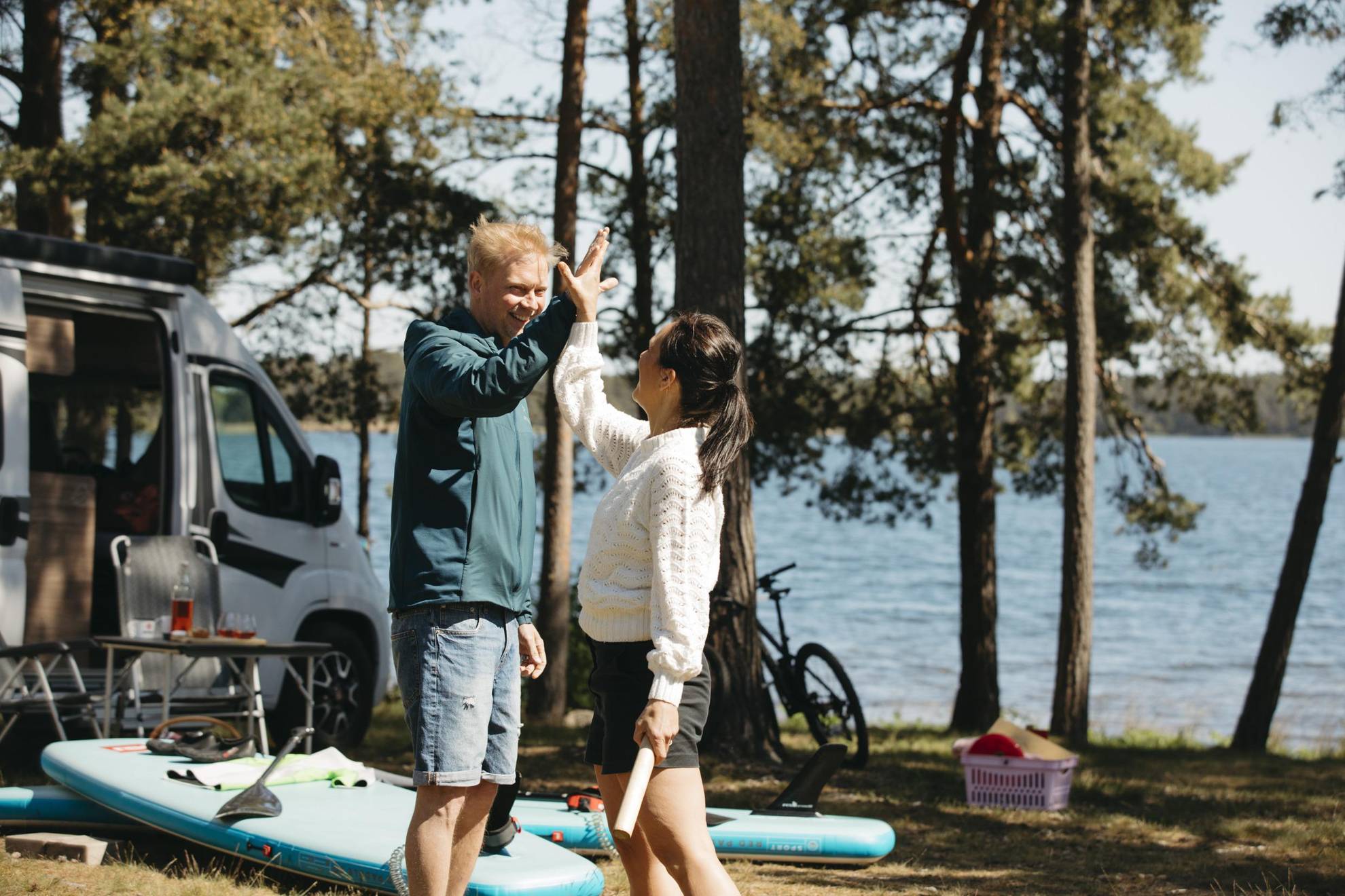 Ein Mann und eine Frau geben sich im Sommer vor ihrem Wohnmobil die Hand. Auf dem Boden liegen Stand Up Paddle Boards.