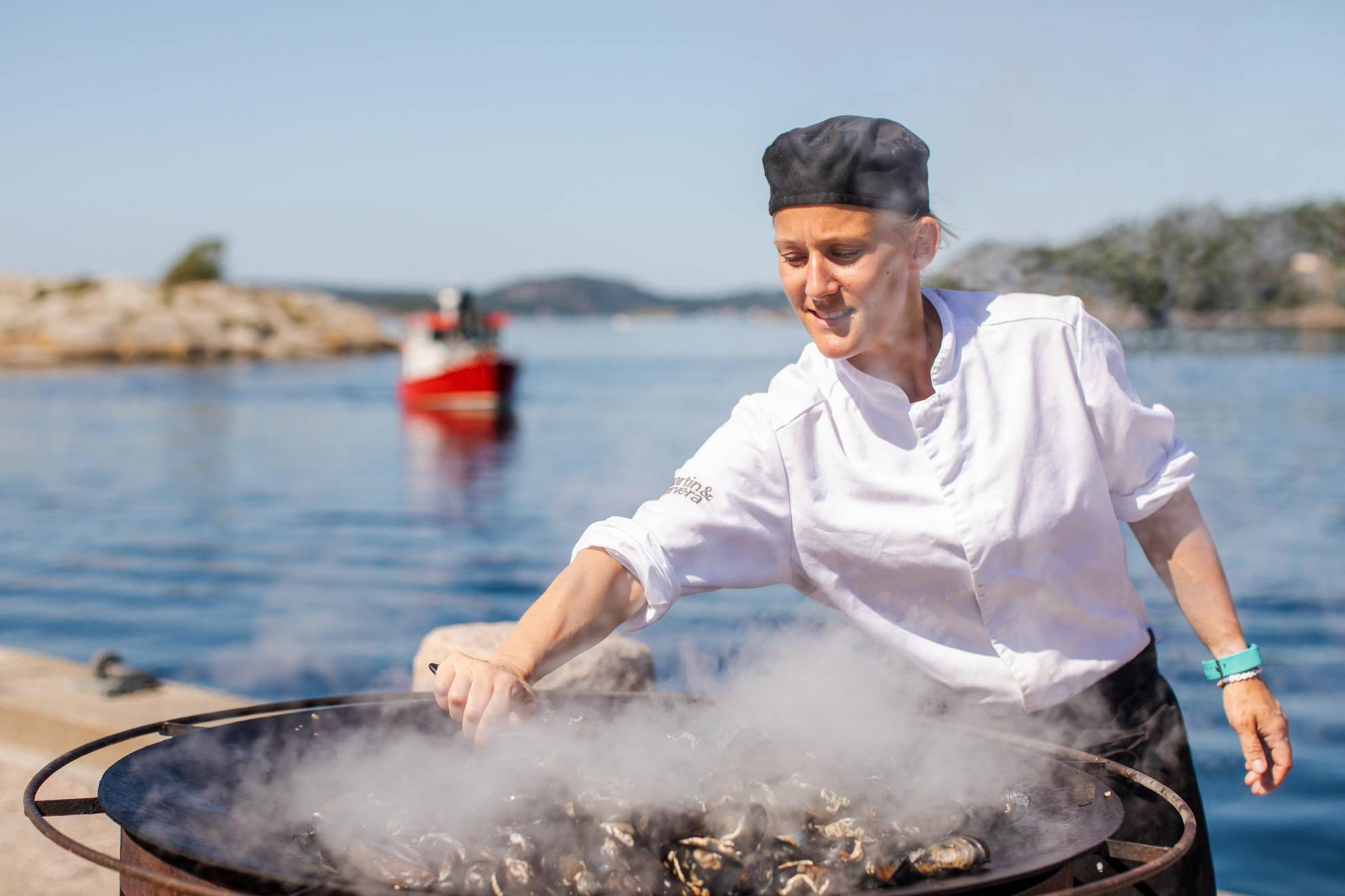 Eine Köchin steht draußen am Meer und kocht Muscheln in einer großen Pfanne.