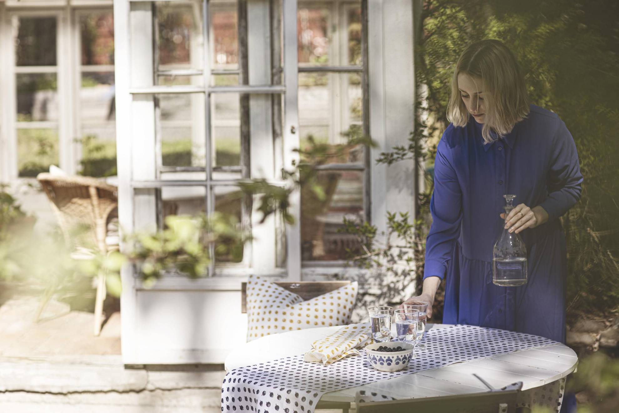 Eine Frau stellt ein Glas Wasser auf einen Tisch, der mit einer Tischdecke und einer Schüssel Blaubeeren gedeckt ist. Im Hintergrund sieht man die Türen zu einem Haus und vorne ist ein Ast verschwommen.