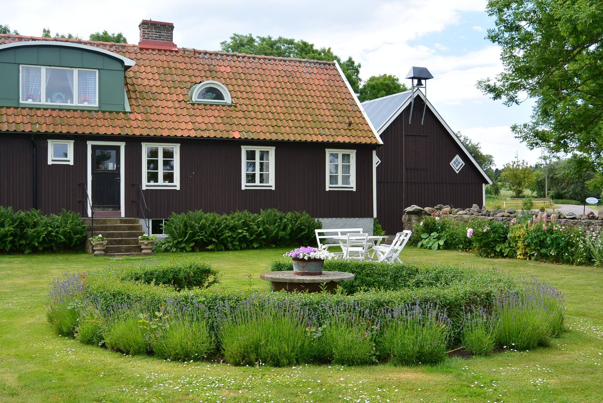 Hinter einem Garten mit Blumenhecke und Sitzecke steht ein großes rotes Haus.