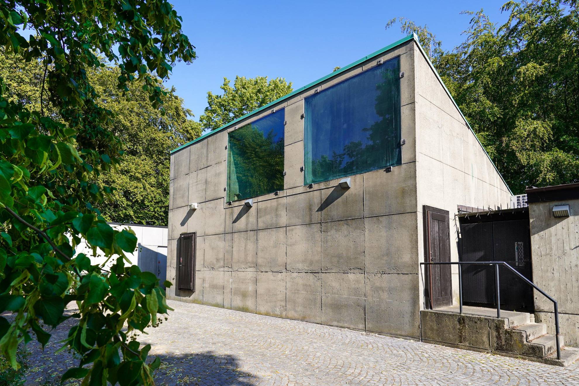Das Haus mit dem Namen "Der Blumenkiosk" ist ein einfaches Betonhaus, dessen Fenster mit schwarzer Dichtungsmasse befestigt sind. Es ist von Grünflächen umgeben.