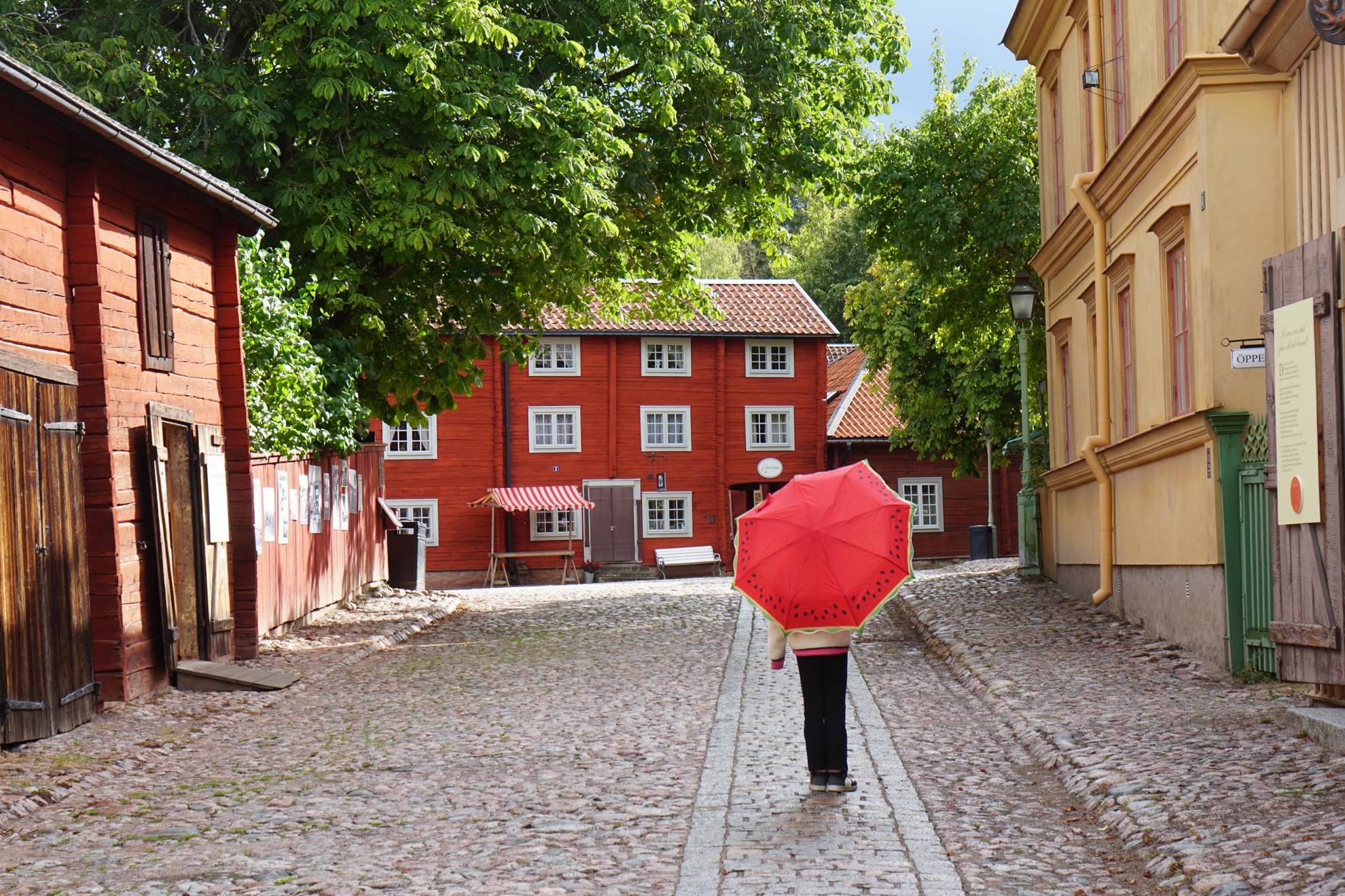 Eine Person geht mit einem roten Regenschirm über einen gepflasterten Weg zwischen alten Häusern