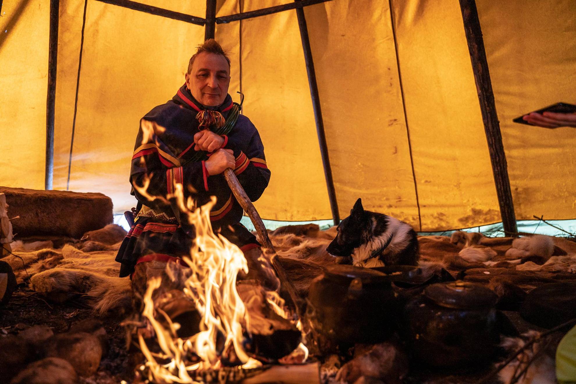 Ein Mann in traditioneller samischer Kleidung sitzt in einem Zelt neben einem Feuer.