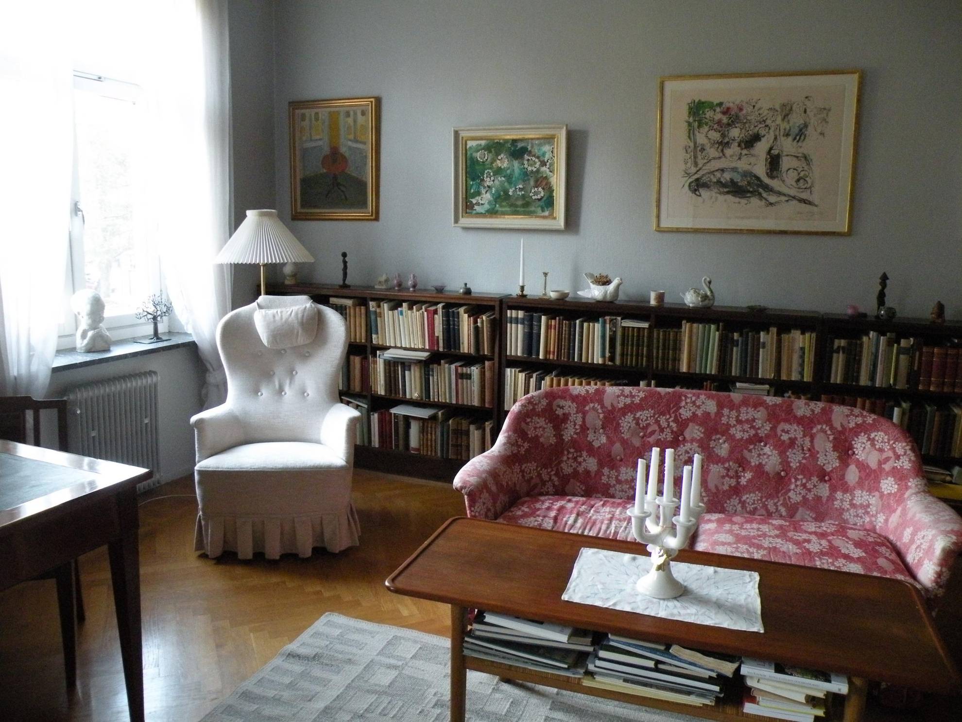 Astrid Lindgren's living room, with a reading chair, a large bookshelf with bibelots on top, a patterned, red couch and a few paintings on the wall.
