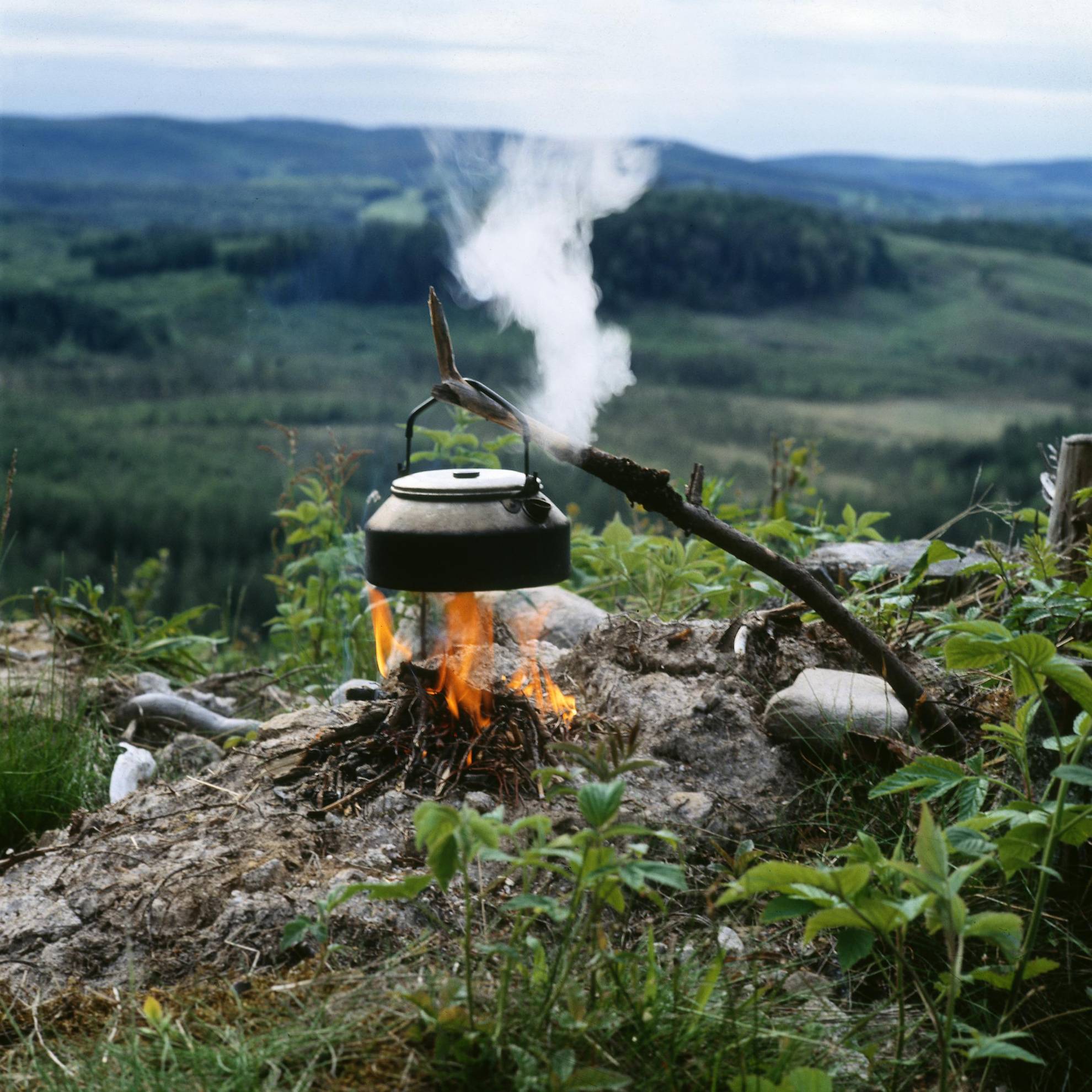 Über einem kleinen Lagerfeuer hängt ein schwarzer Topf an einem Stock. Es dampft aus dem Topf. Im Hintergrund öffnet sich ein Tal mit  bewaldeten Hügeln.