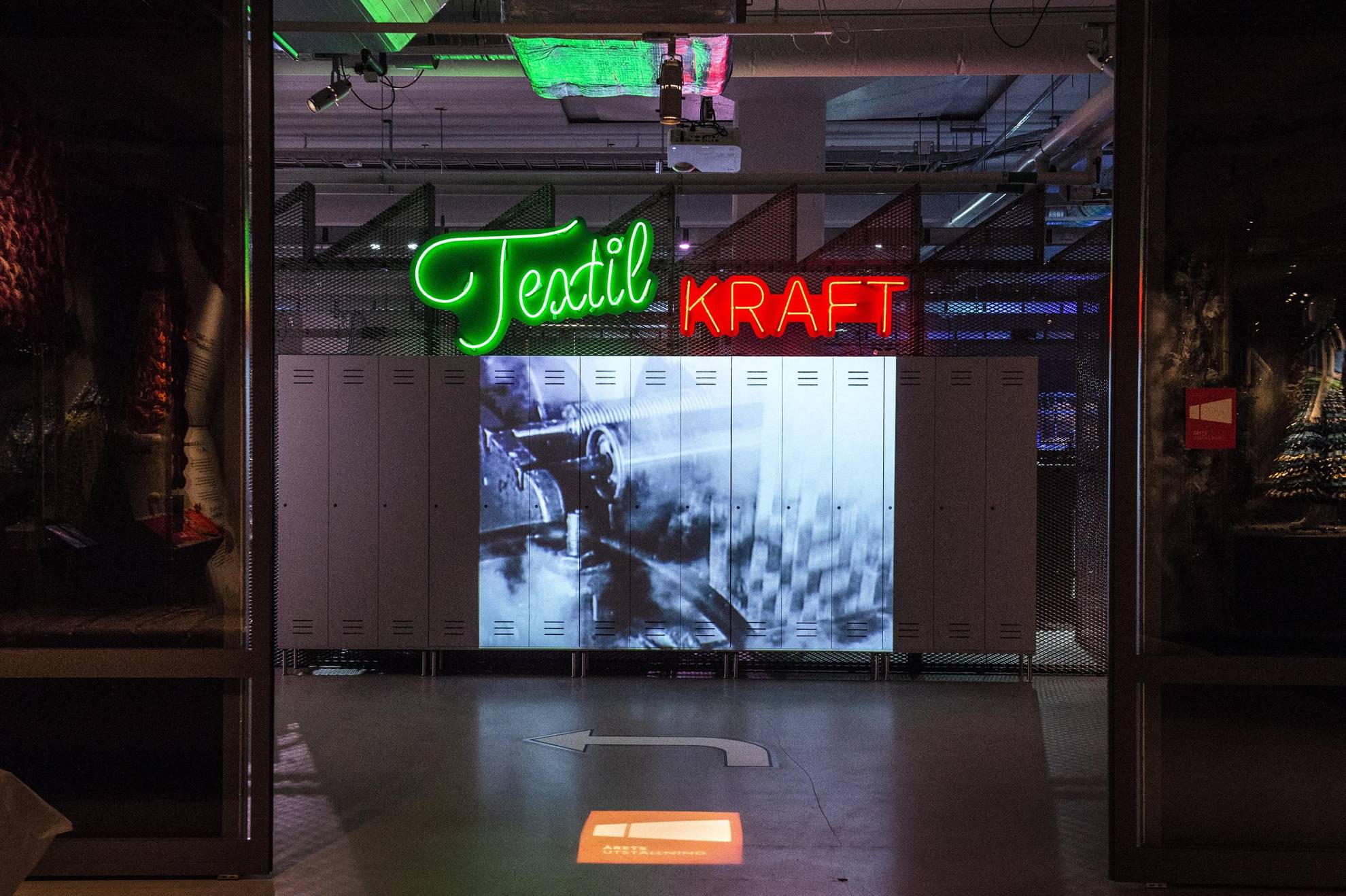 Ein Schwarz-Weiß-Foto einer Nähmaschine, das auf eine Wand im Textilmuseum von Schweden projiziert wird. In grüner Neonschrift darüber das Wort "Textil", in roter Neon-Schrift das Wort "Kraft".