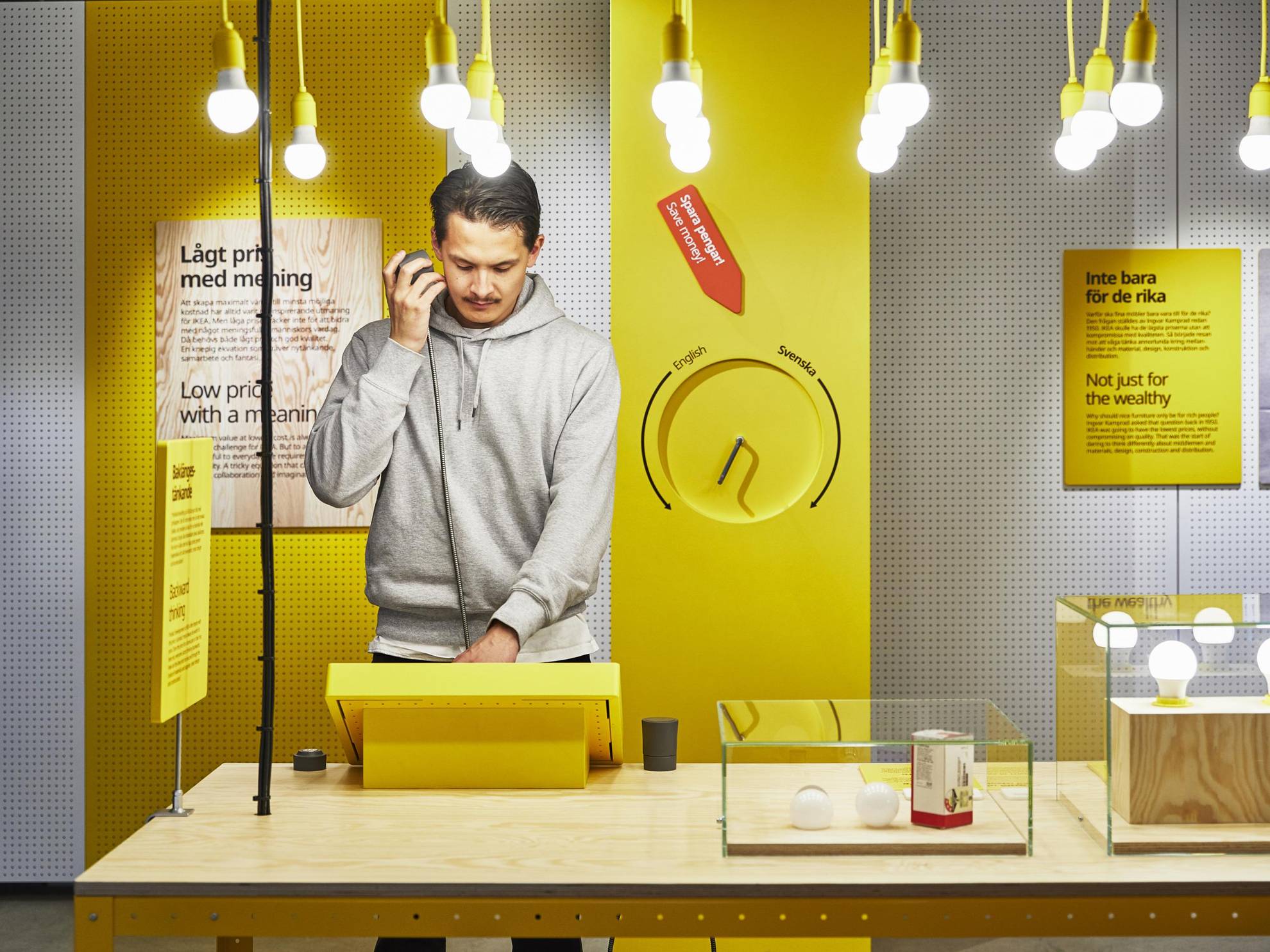 Ein Mann steht an einem Tisch und betrachtet ein gelbes Display in einer der Ausstellungen von Ikea. Von der Decke hängen Glühbirnen und auf dem Tisch Glaskästen mit Glühbirnen drin.