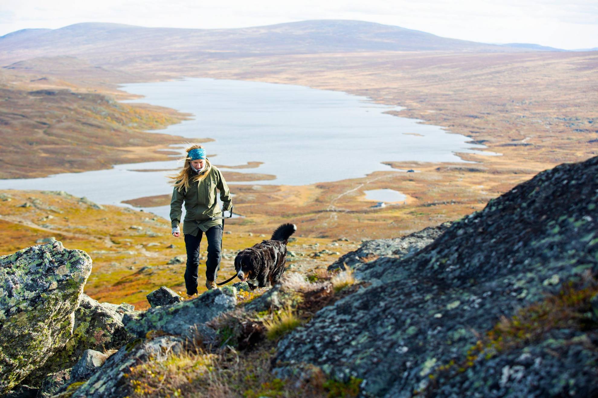 Eine Frau wandert mit ihrem Hund auf einen felsigen Berg. Im  Hintergrund ein breiter Fluss in einer herbstlichen Landschaft.
