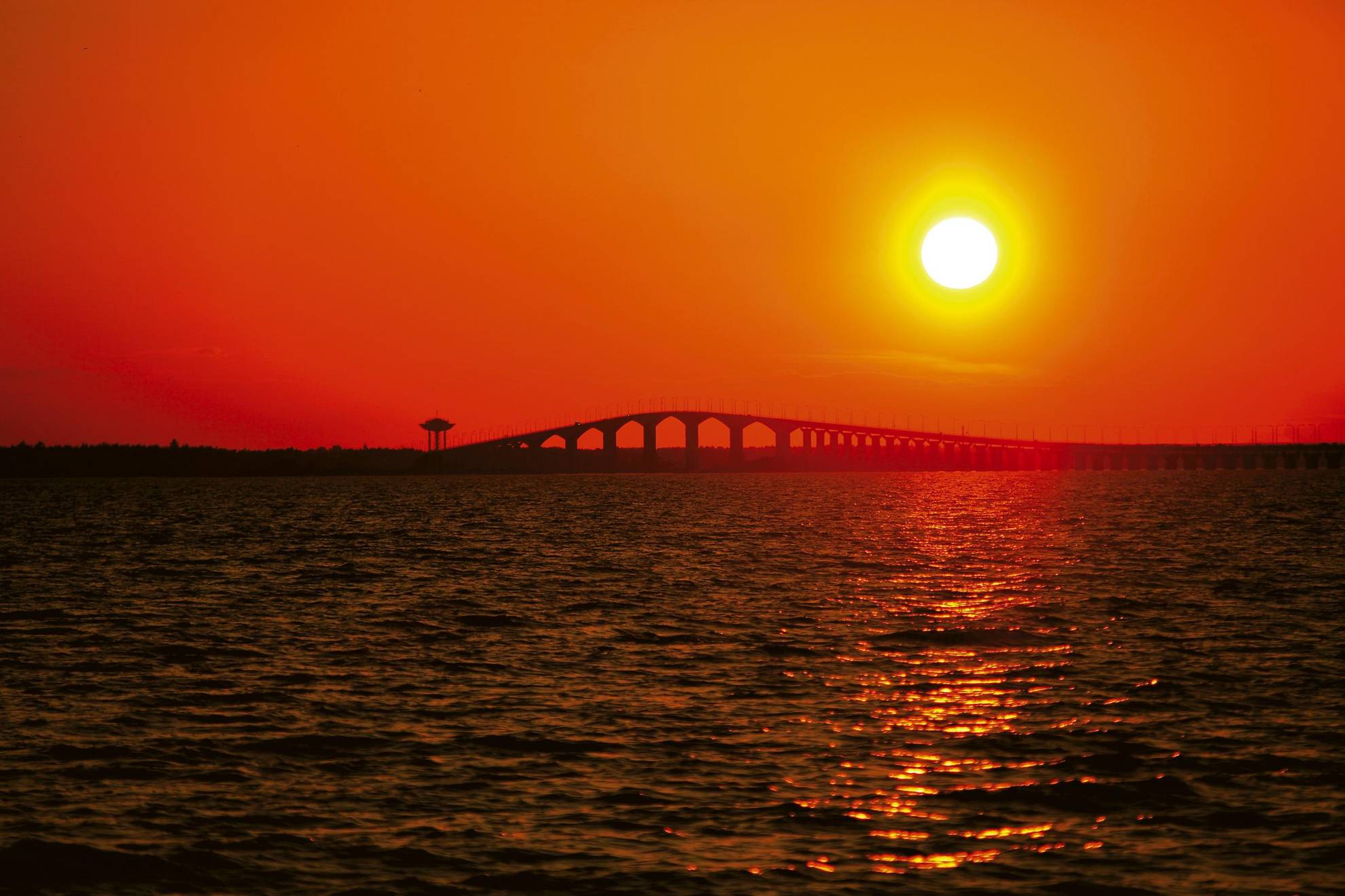 Die Sonne geht über der Brücke Ölandsbron unter. Der Himmel ist orange und die gelbe Sonne macht eine Solarstraße im Wasser.