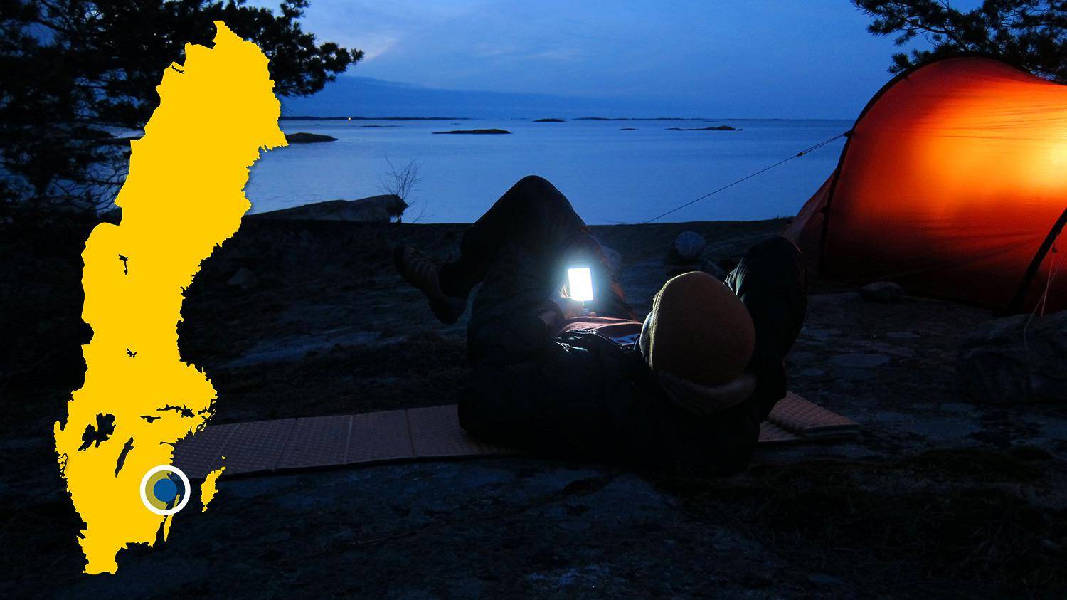 An einem Strand in der Nähe des Wassers wird ein Zelt aufgebaut. Es ist Nacht und eine Person liegt neben dem Zelt und schaut auf ihr Handy.