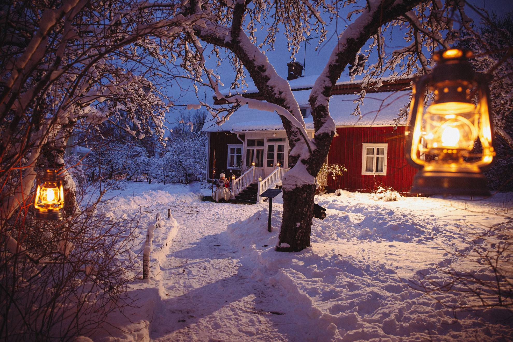 Ein rotes Häuschen mit weißen Ästen, umgeben von Schnee. Eine Frau sitzt auf den Stufen des Hauses.