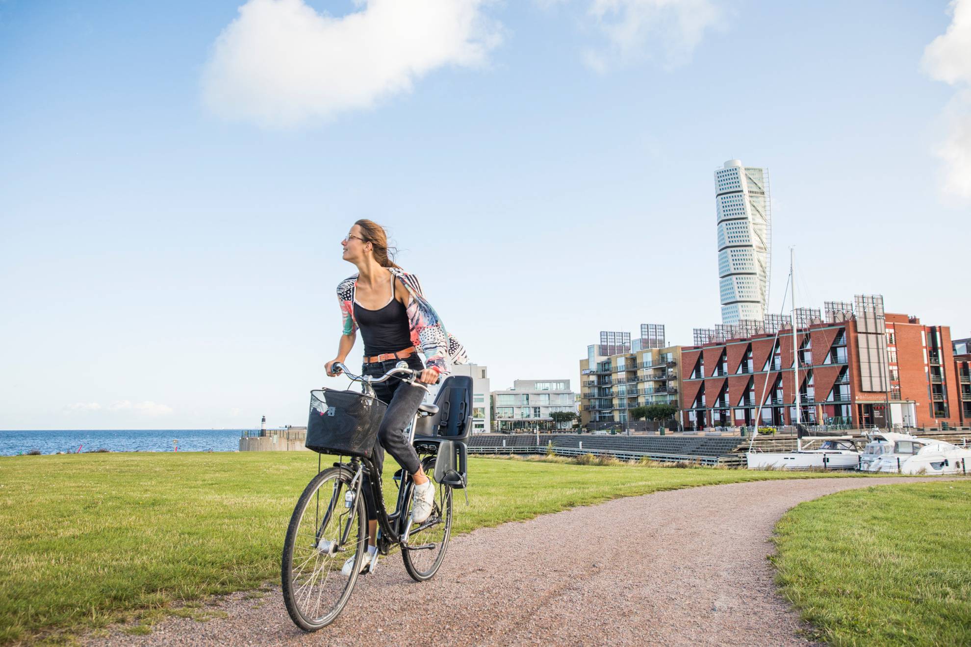 Eine Frau fährt mit dem Fahrrad auf einem Gehweg, dahinter siehst du ein Wohngebiet am Meer und in der Mitte einen Wolkenkratzer.