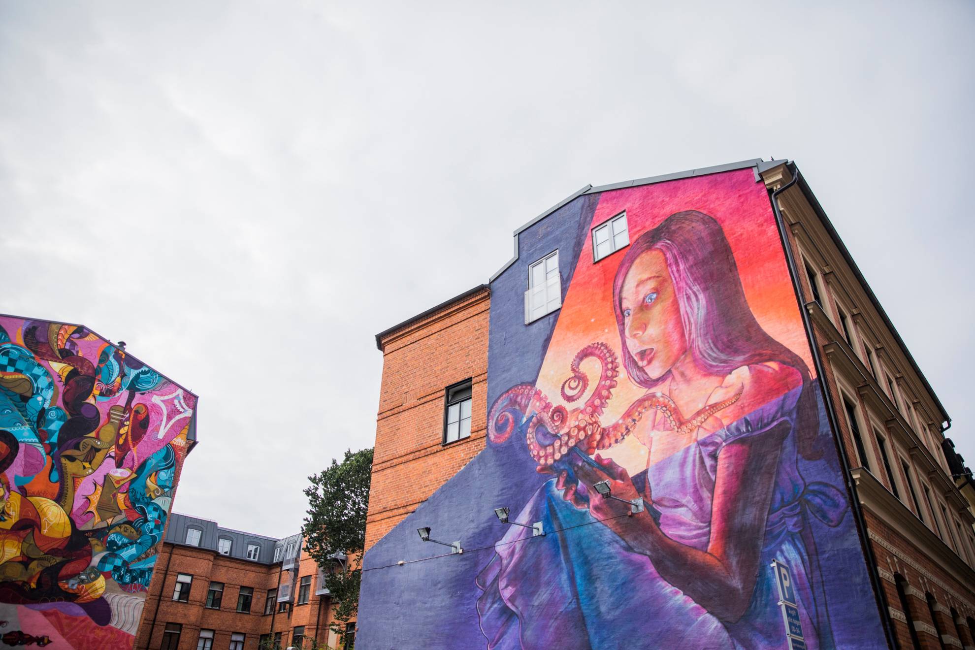 Ein Wandgemälde an einem Haus in Malmö, das eine Frau zeigt, die einen Oktopus hält, und ein Haus daneben, auf dem Teile der abstrakten Malerei zu sehen sind.