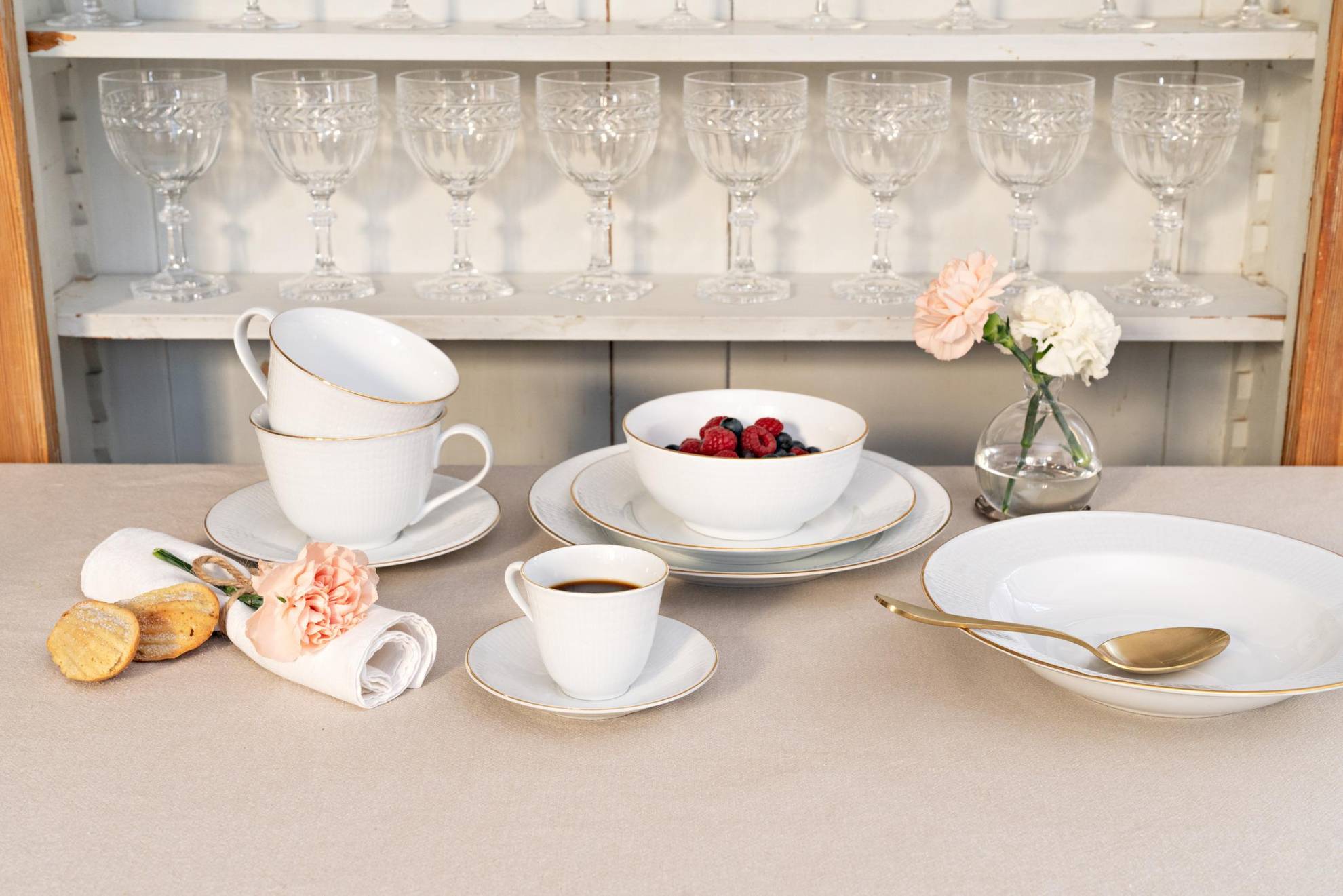 Ein Geschirrservice namens „Swedish Grace“ auf einem Tisch. Teller in verschiedenen Größen eine Schale mit Beeren, zwei Teetassen und eine Tasse Kaffee. Dahinter befindet sich ein Regal mit mehreren Weingläsern.