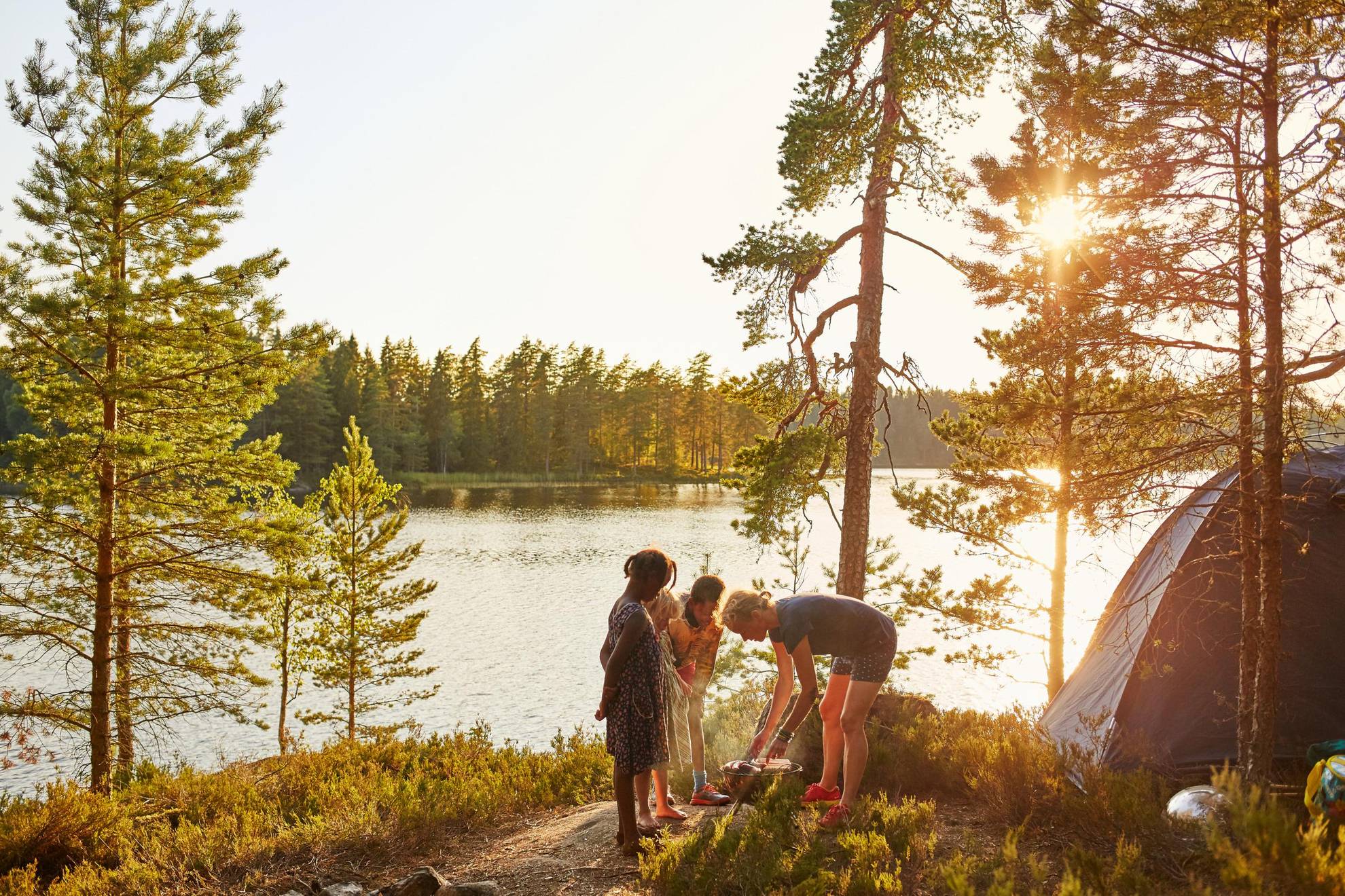 : People in Sweden barbecuing by their tent, next to a lake in the forest.