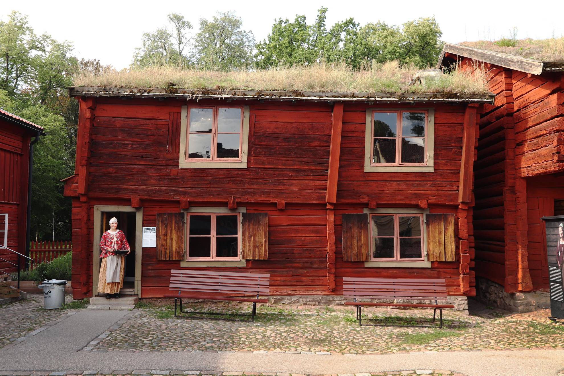 Ein altes rot gestrichenes Holzhaus mit Grasdach. Eine Frau steht am Eingang des Hauses.