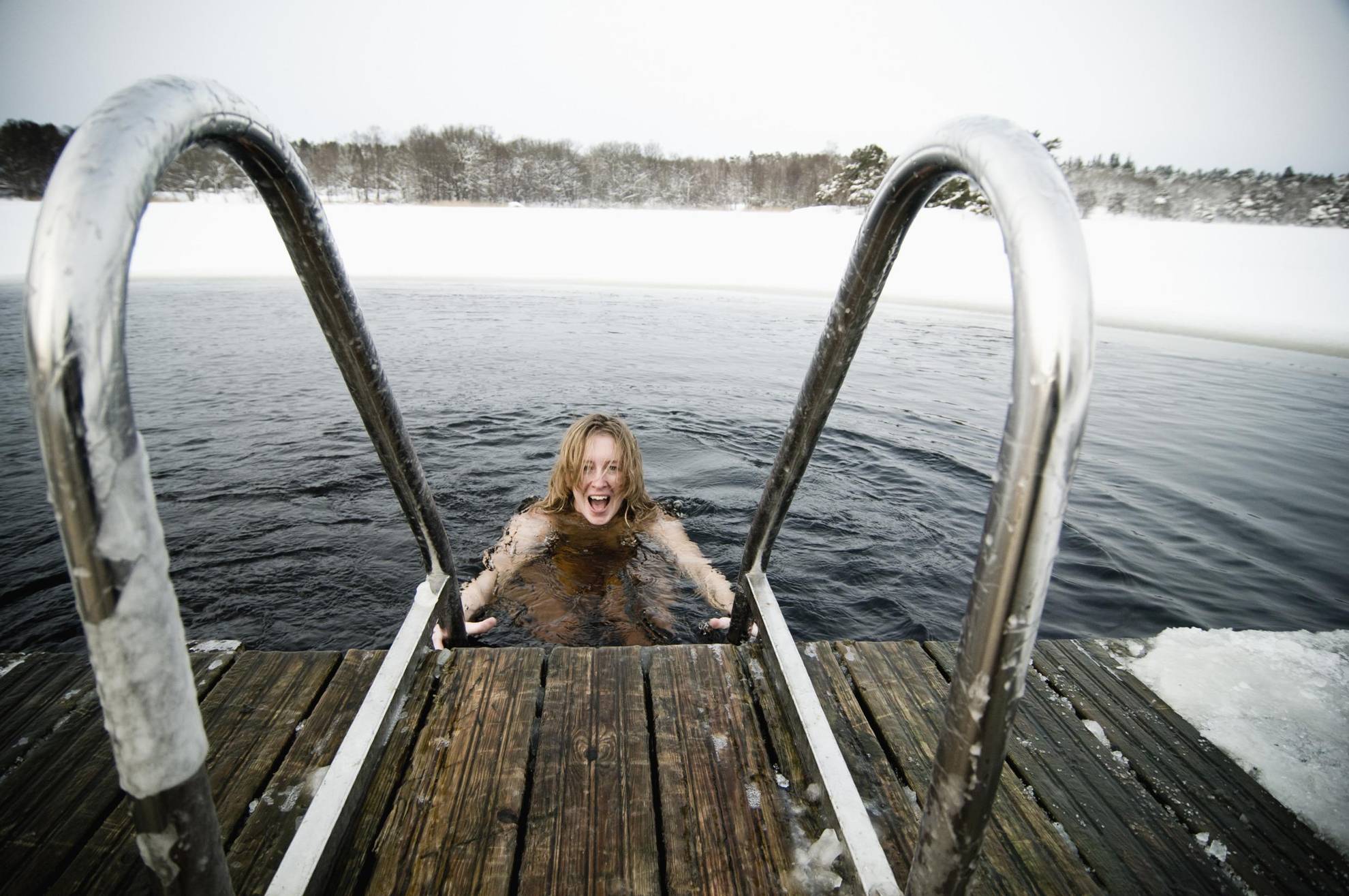 Eine Frau will aus dem See eine Treppe zum Holzsteg hinaussteigen. Der See ist mit Schnee und Eis bedeckt mit Ausnahme der Ausstigesstelle.