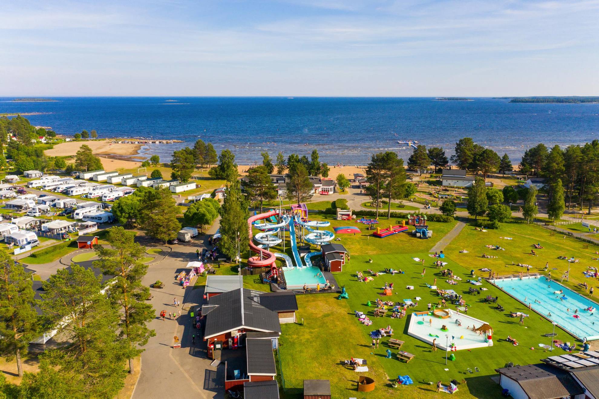 Eine Luftaufnahme von Byske Camping während des Sommers mit Stellplätzen links, einem Schwimmbad mit Wasserrutsche rechts und dem Meer im Hintergrund.