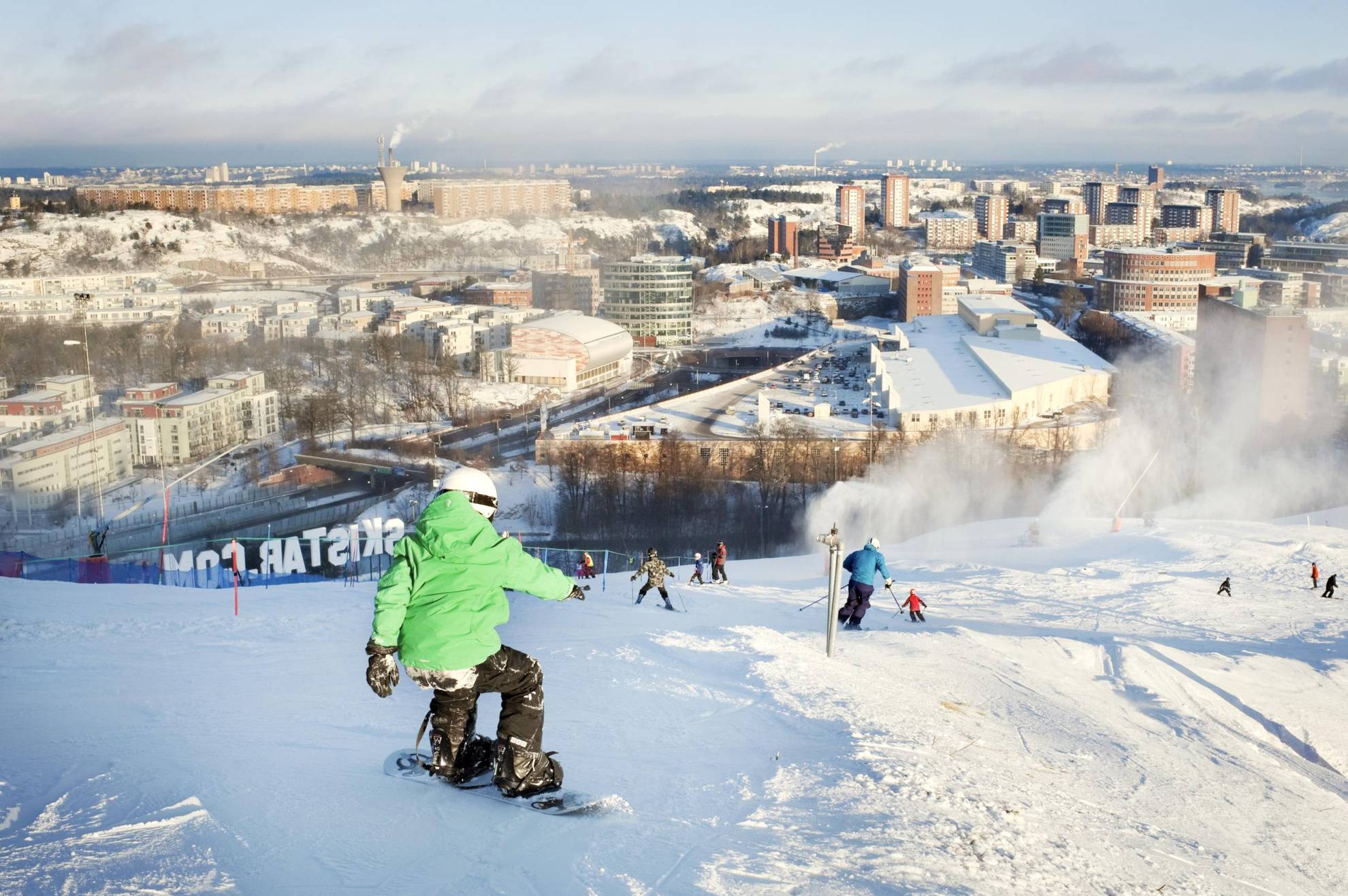 Leute fahren auf Skiern und Snowboards die Pisten von Hammarbybacken hinunter und blicken über die Innenstadt von Stockholm.
