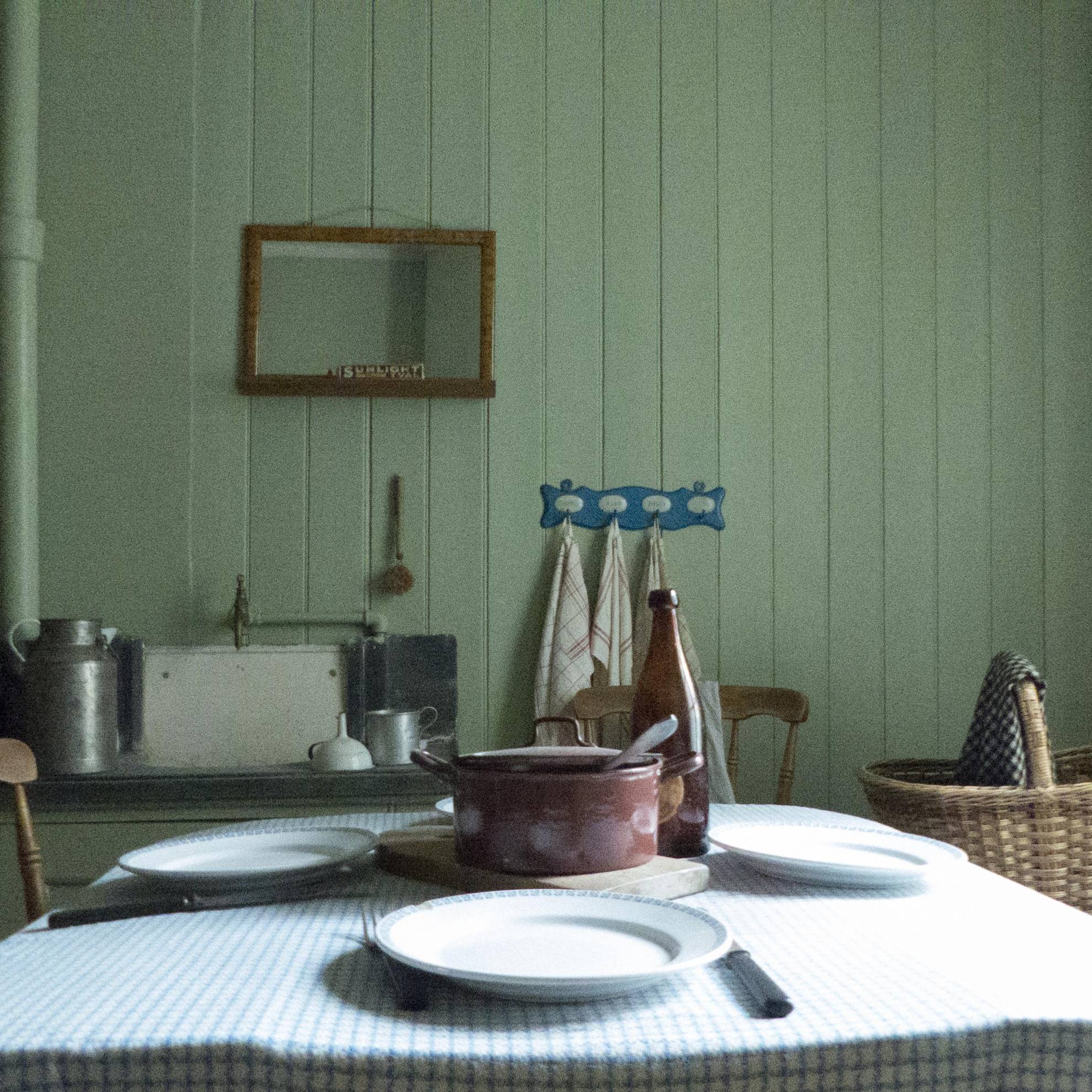 In einem Arbeiterheim im ersten Vorort von Göteborg im 19. und 20. Jahrhundert. Ein gedeckter Tisch mit einem Eisentopf in der Mitte. An einer grünen Holzwand hängt ein Spiegel.
