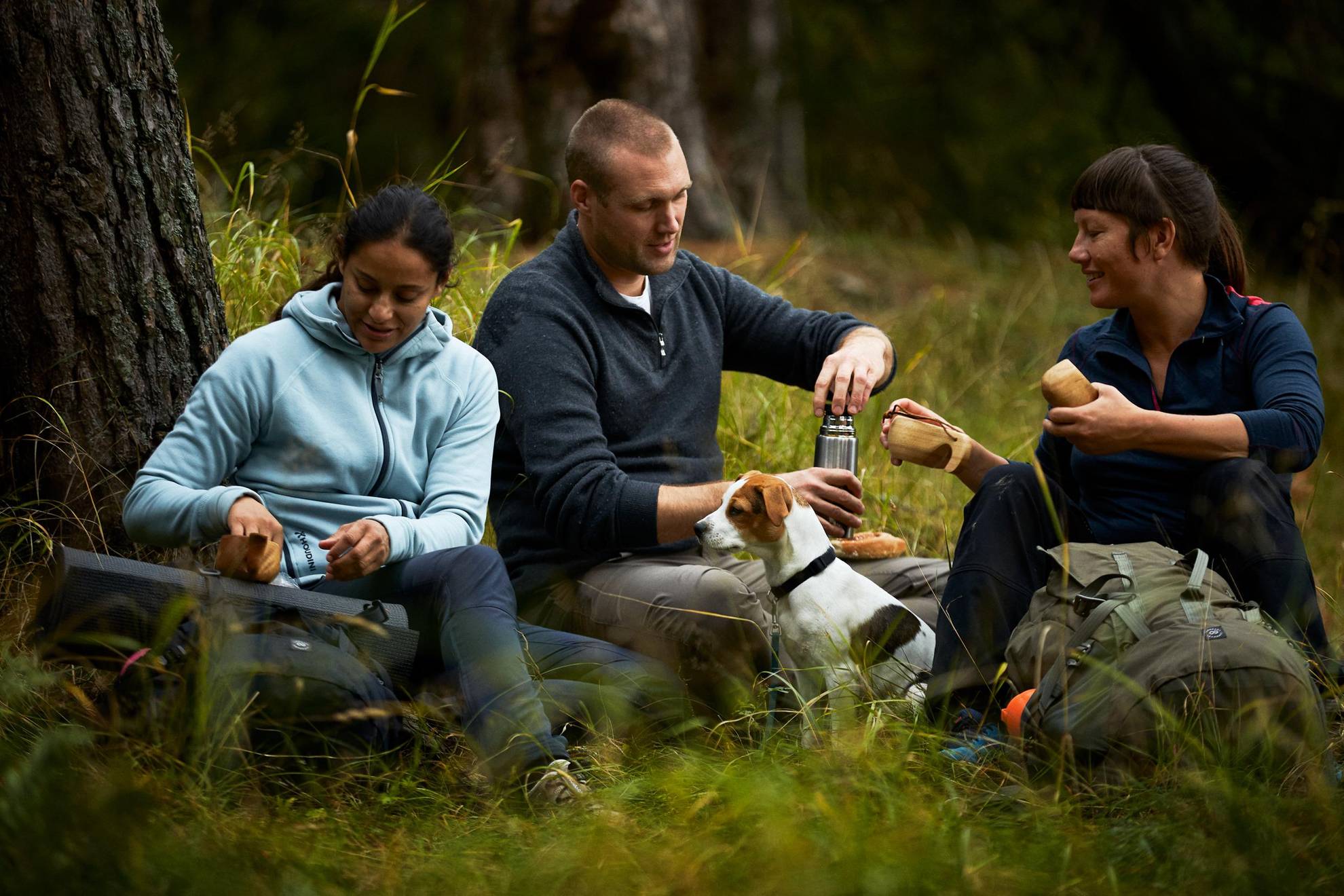 Wanderer genießen eine Fika im Freien. Sie sitzen im Grünen und trinken aus Holzbechern ein Heißgetränk und essen Zimtschnecken.
