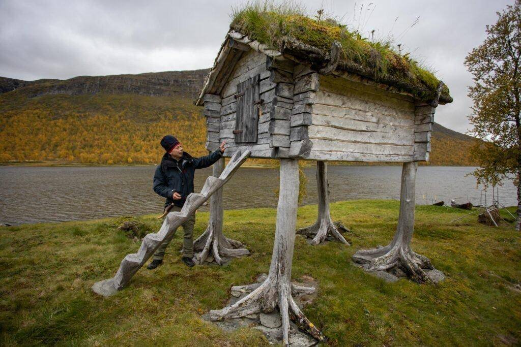 Eine Person steht neben einem kleinen Holzhaus, das auf Baumstämmen aufgestellt ist. Im Hintergrund ein Fluss.