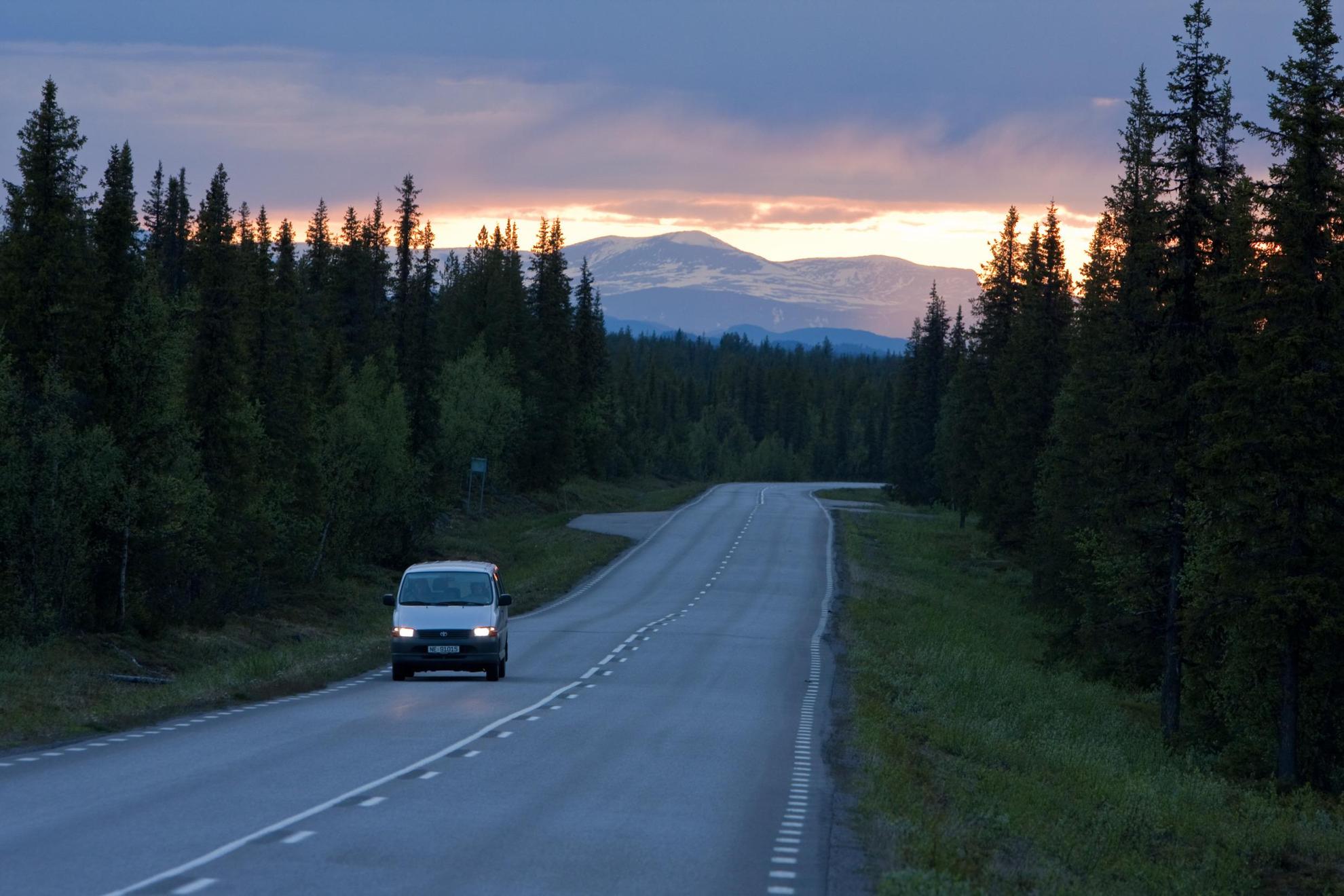 A minivan is driving along a road during the midnight sun in northern Sweden, forests on both sides and mountains in the background.