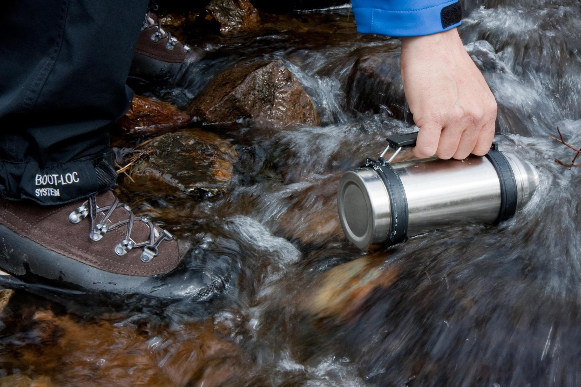 Someone fills a metal flask with water from a stream in the mountains.