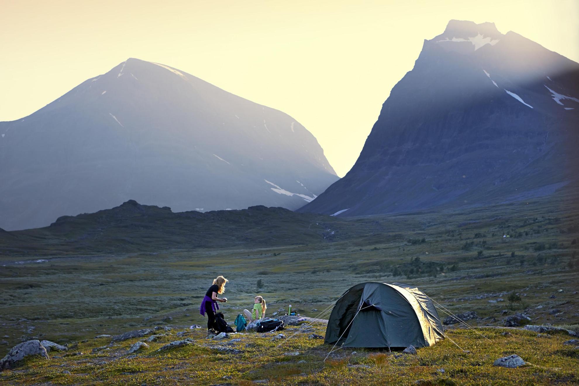 Eine Frau sitzt mit ihrer Tochter neben einem aufgebauten Zelt. Hohe Berge im Hintergrund.
