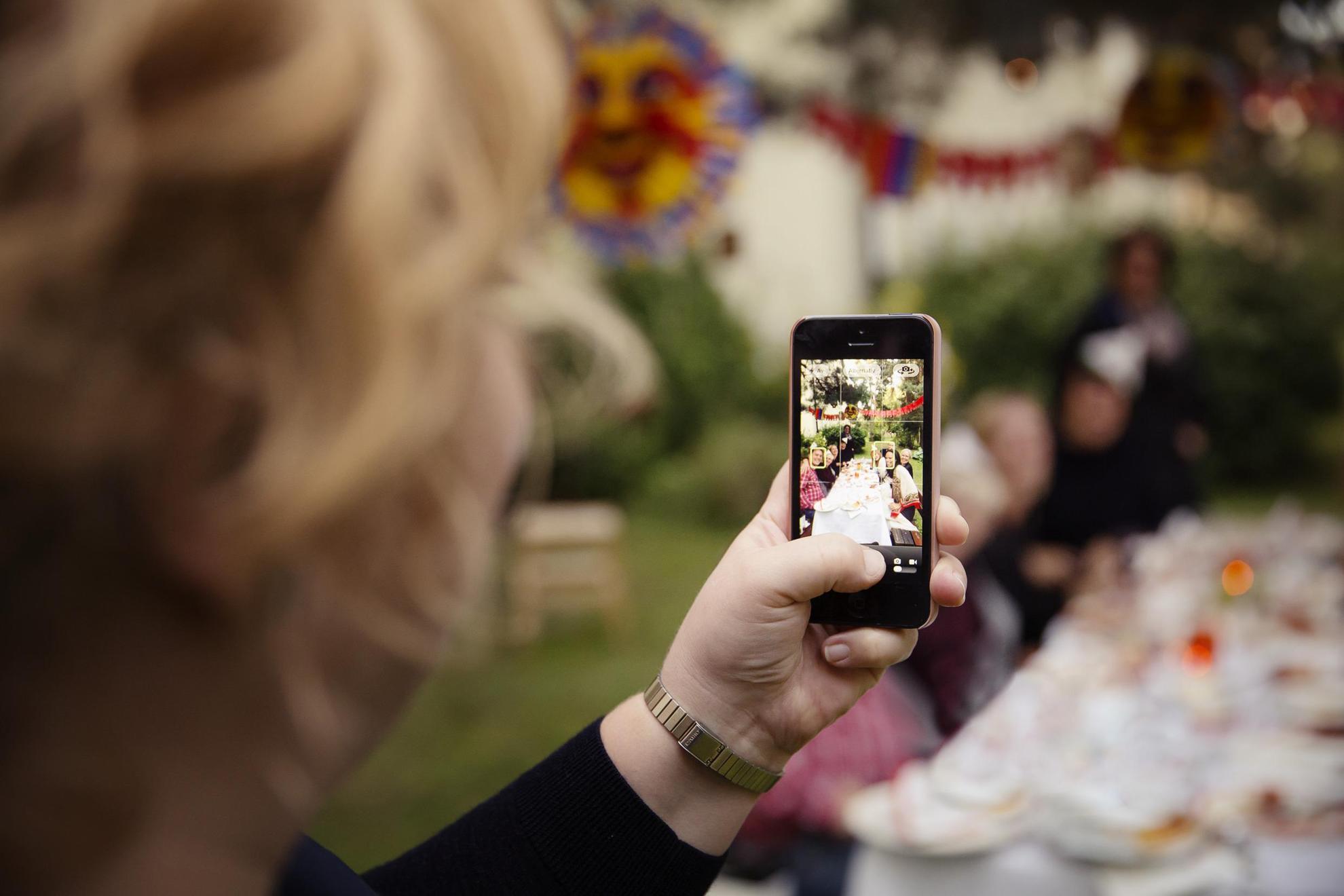 A woman is holding up a phone, taking pictures of the people sitting down by the decorated party table.