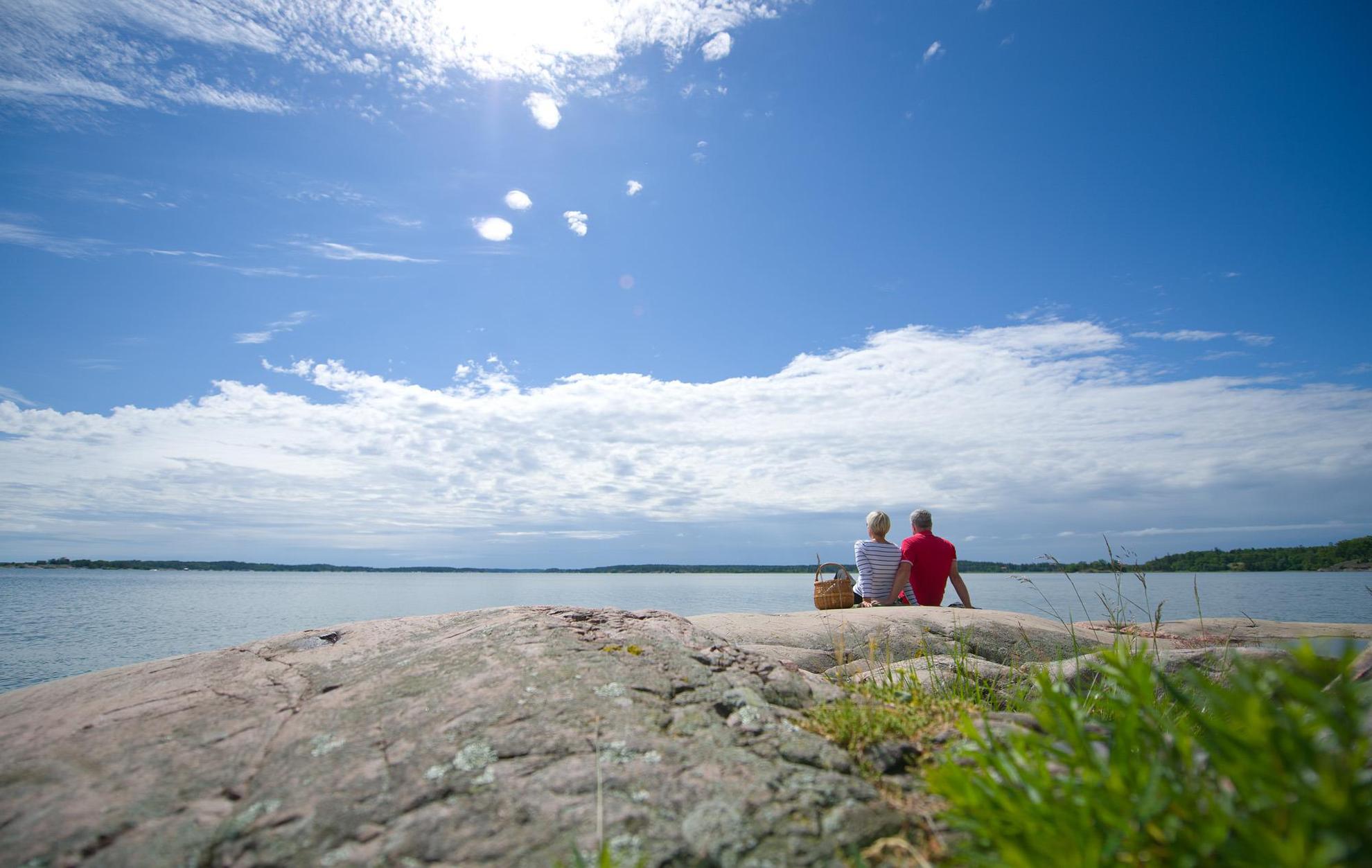 Ein Paar mit Picknickkorb sitzt nebeneinander auf einem Felsen am Meer.