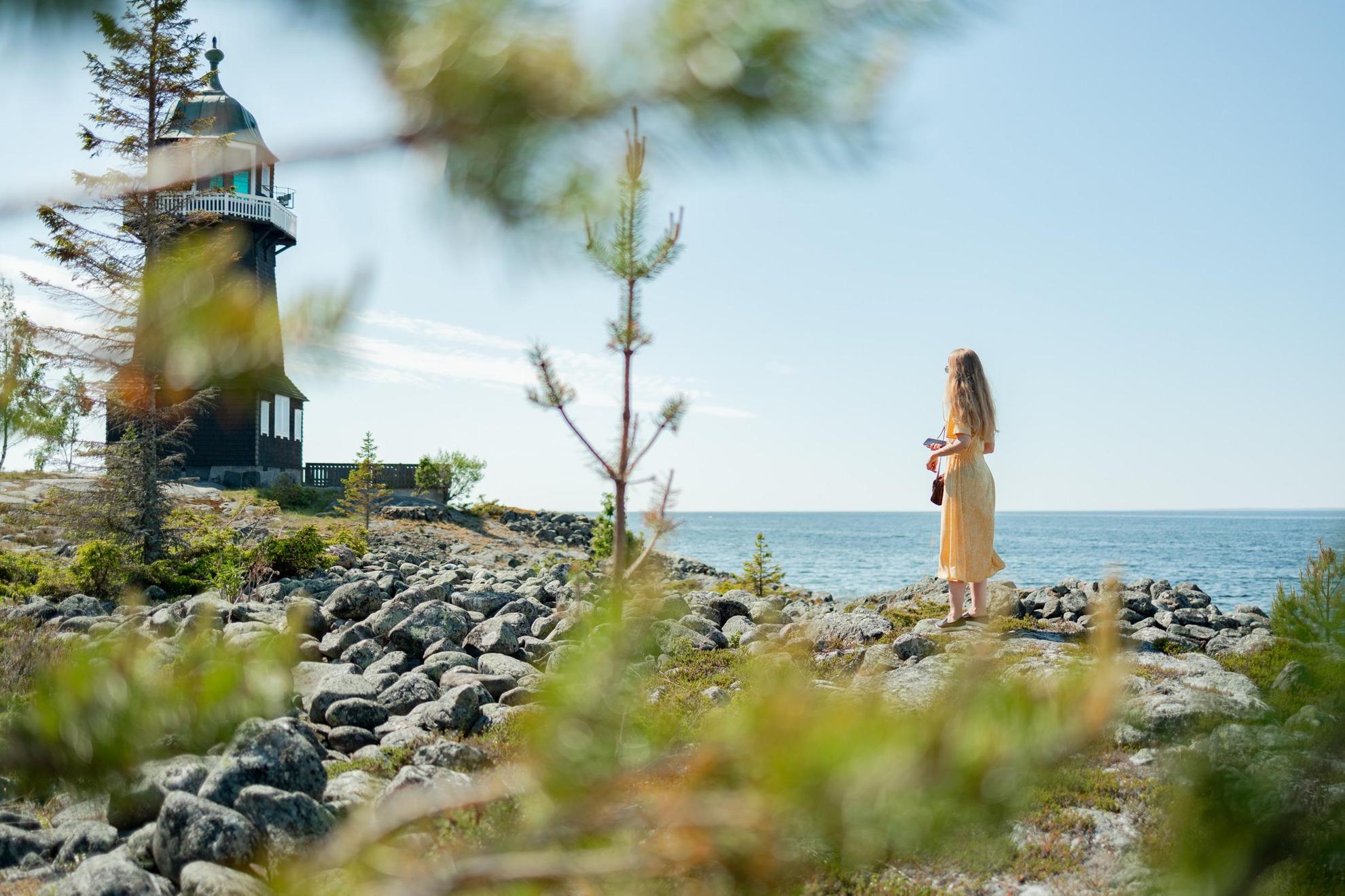 Eine Frau in einem gelben Kleid blickt auf einen roten Leuchtturm am Meer.