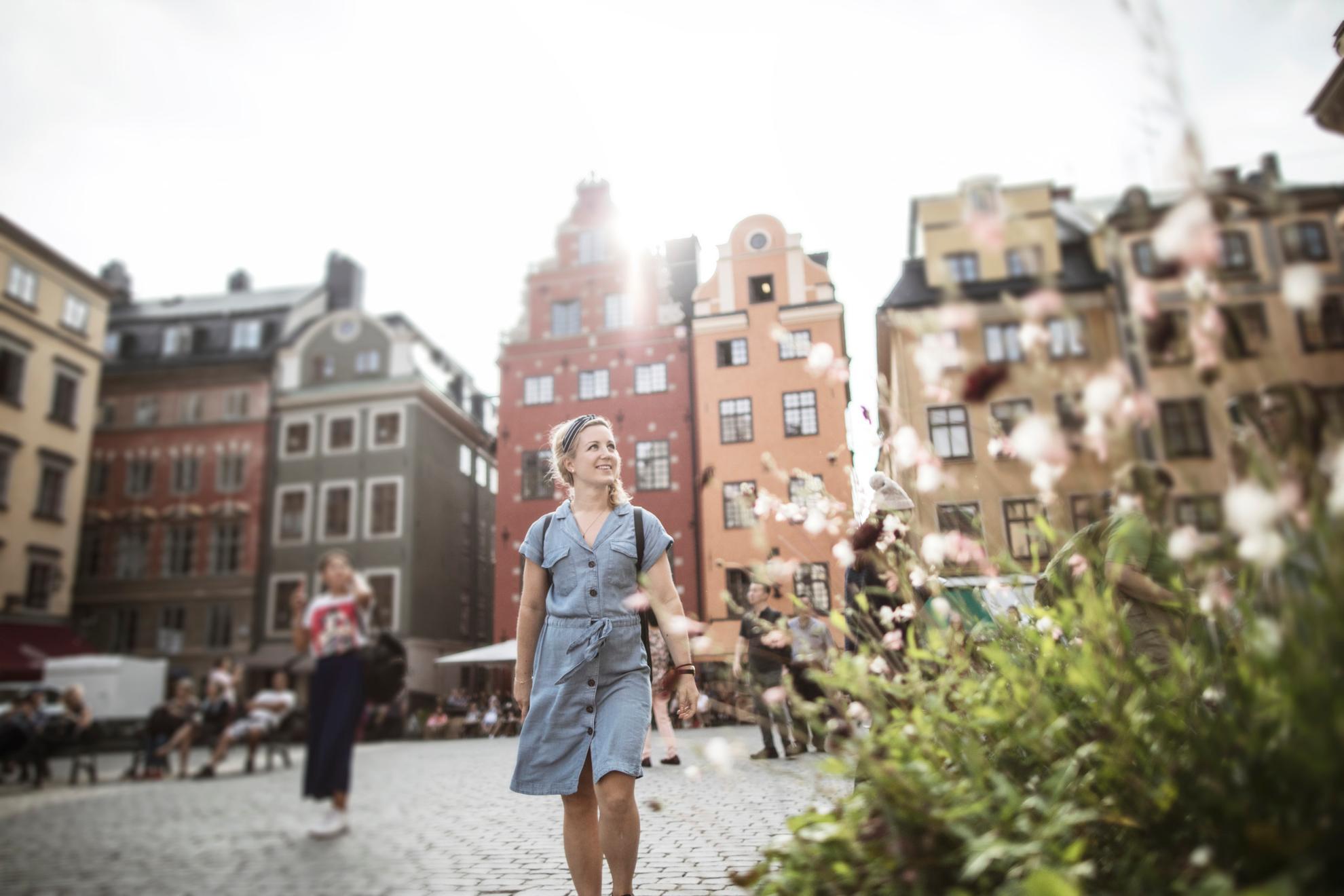 Eine Frau geht in Stockholm spazieren. Im Hintergrund siehst du alte Steinhäuser und im Vordergrund steht eine blühende Pflanze.