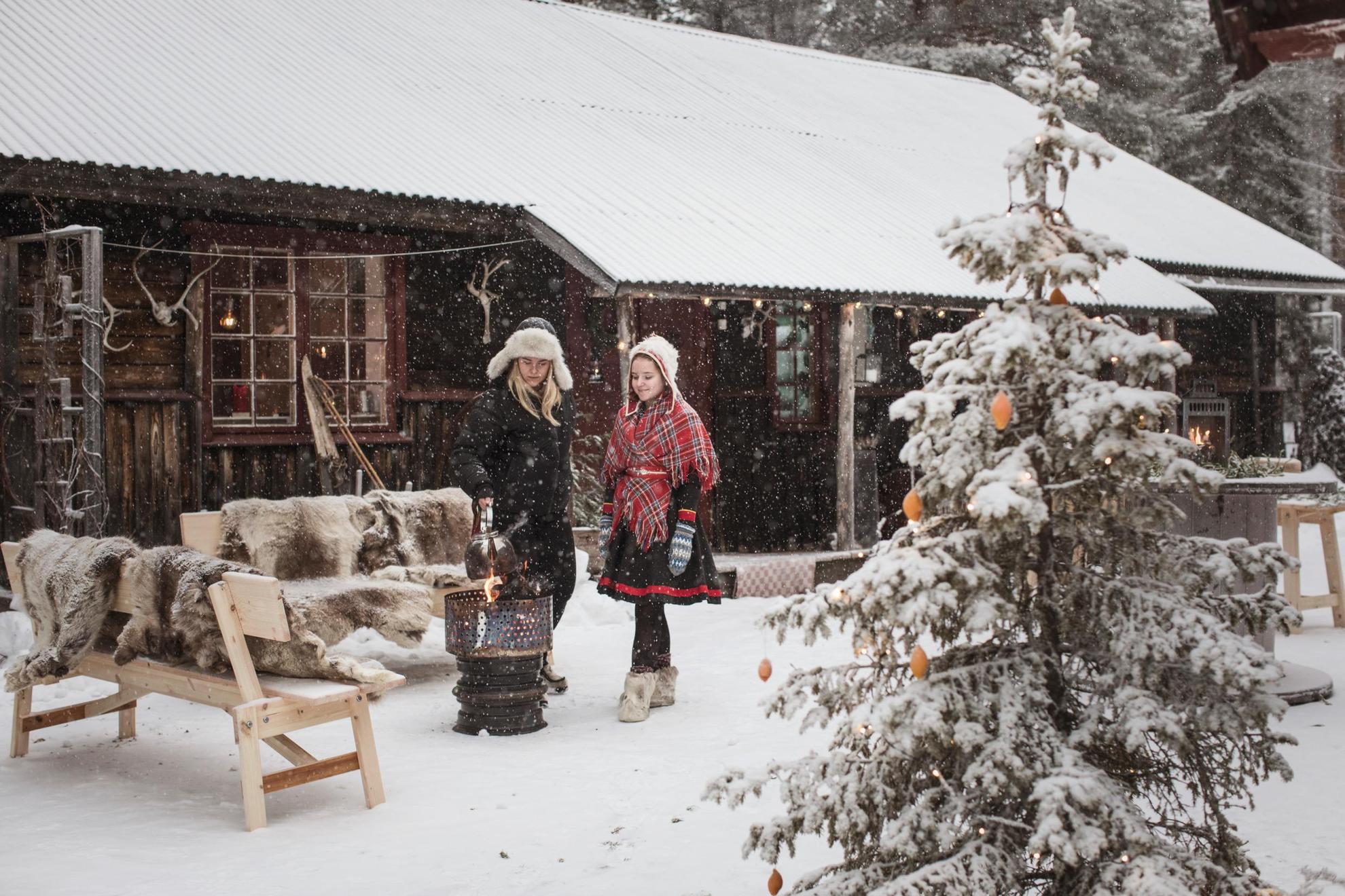 Zwei junge Frauen stehen im Schnee vor einem Holzhaus. Eines der Mädchen trägt eine samische Tracht. Sie kochen über einem offenen Feuer. Auf den Bänken liegen Rentierfelle und vorne steht eine Tanne.