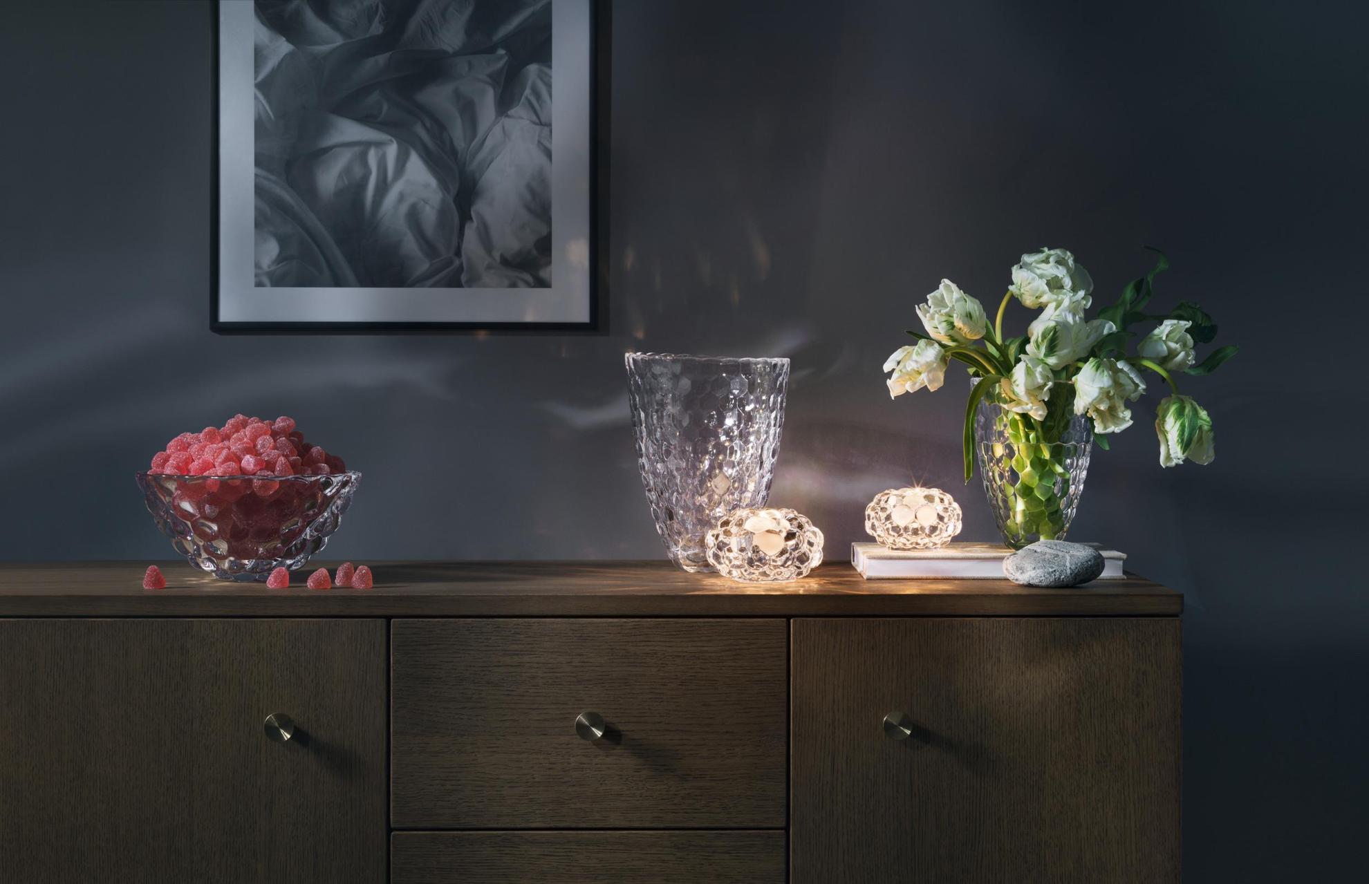 Designobjekte aus Glas auf einer Holzkommode, zwei Kerzenhalter mit brennenden Kerzen, zwei Vasen, eine mit Tulpen und eine mit roten Bonbons gefüllte Schale.