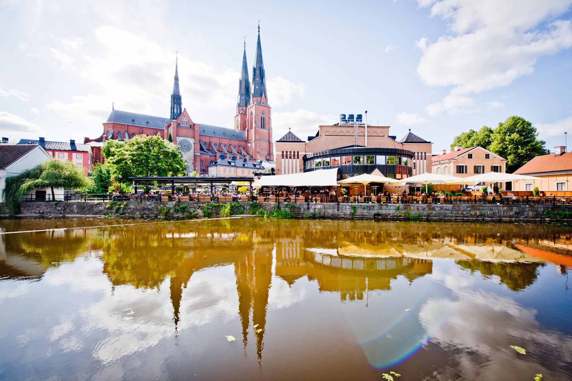 Der Dom von Uppsala ragt im Hintergrund in den Himmel. Davor stehen andere Gebäude und eine Außenterrasse mit vielen Stühlen. Im Wasser des Fluss Fyris, spiegeln sich die Gebäude der Stadt.