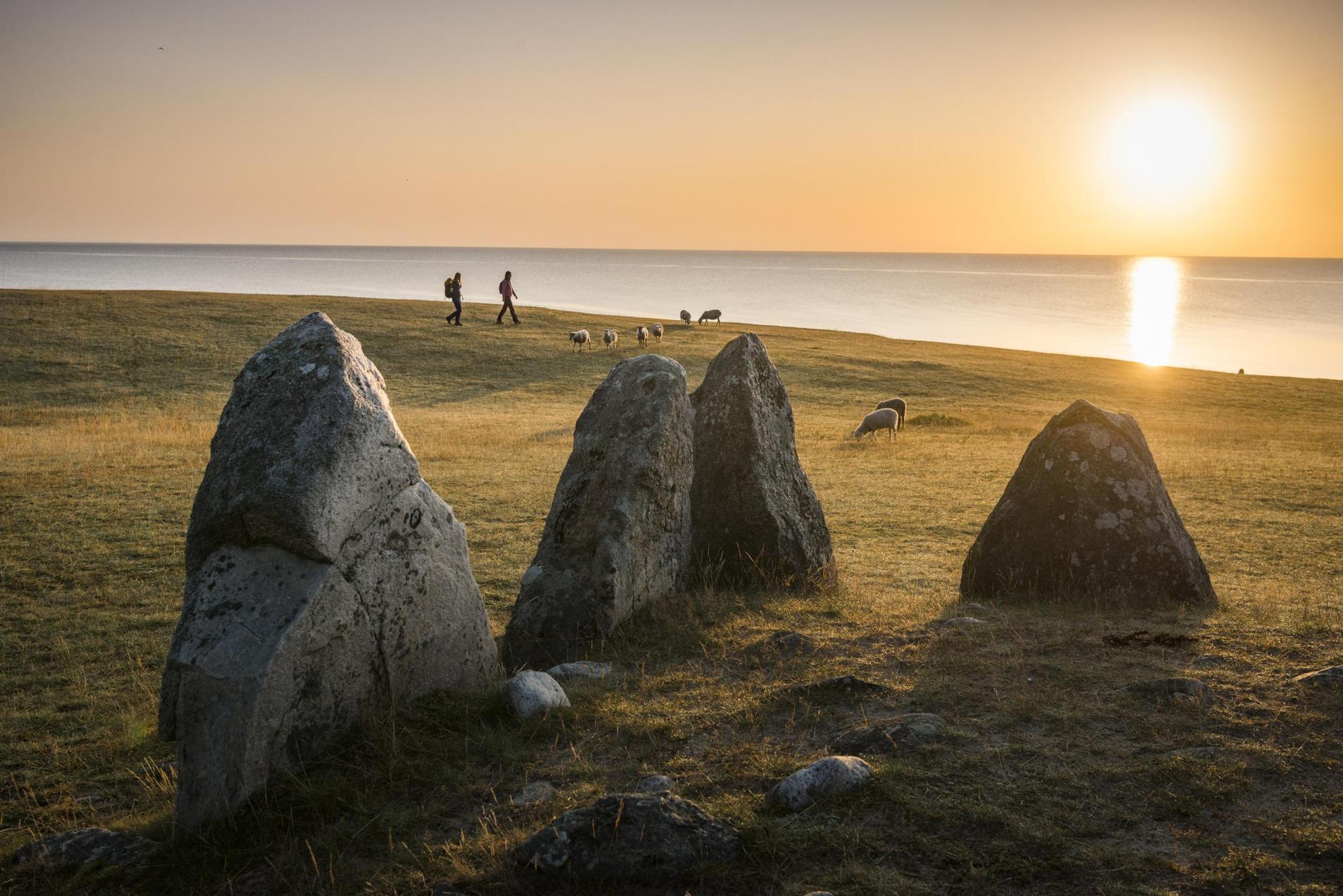 Ein megalithisches Monument in Form eines Steinschiffs. 4 der 59 Felsblöcken sind auf dem Bild zu erkennen. Him Hintergrund spazieren Wanderer.Schafe grassen auf der Wiese. Die Sonne spiegelt sich im Meerwasser.