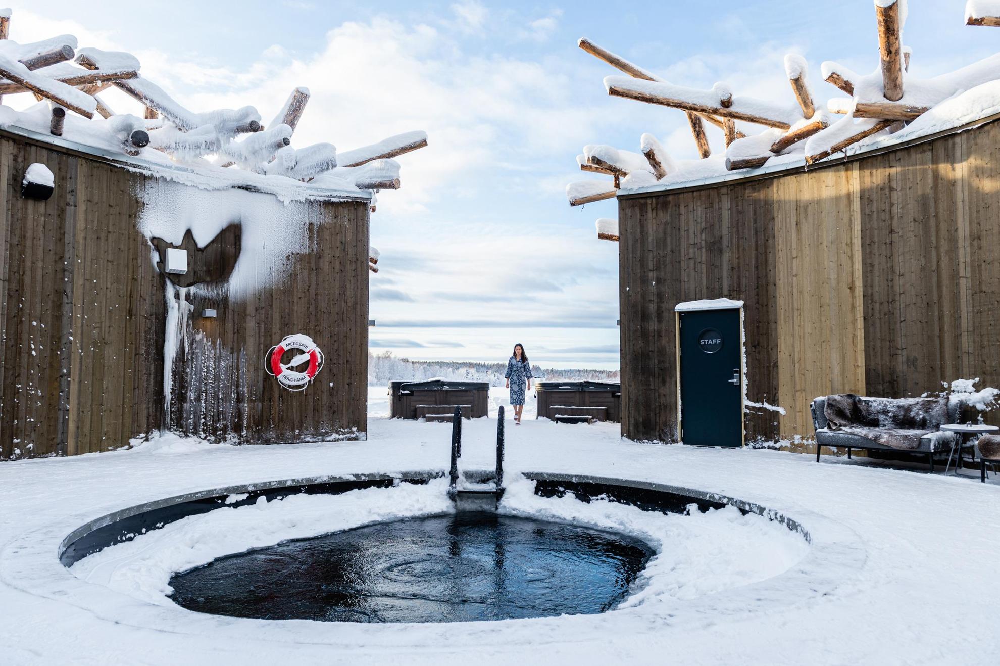 Eine Frau geht auf ein offenes Bad im Schnee zu, das von einem Holzgebäude umgeben ist.