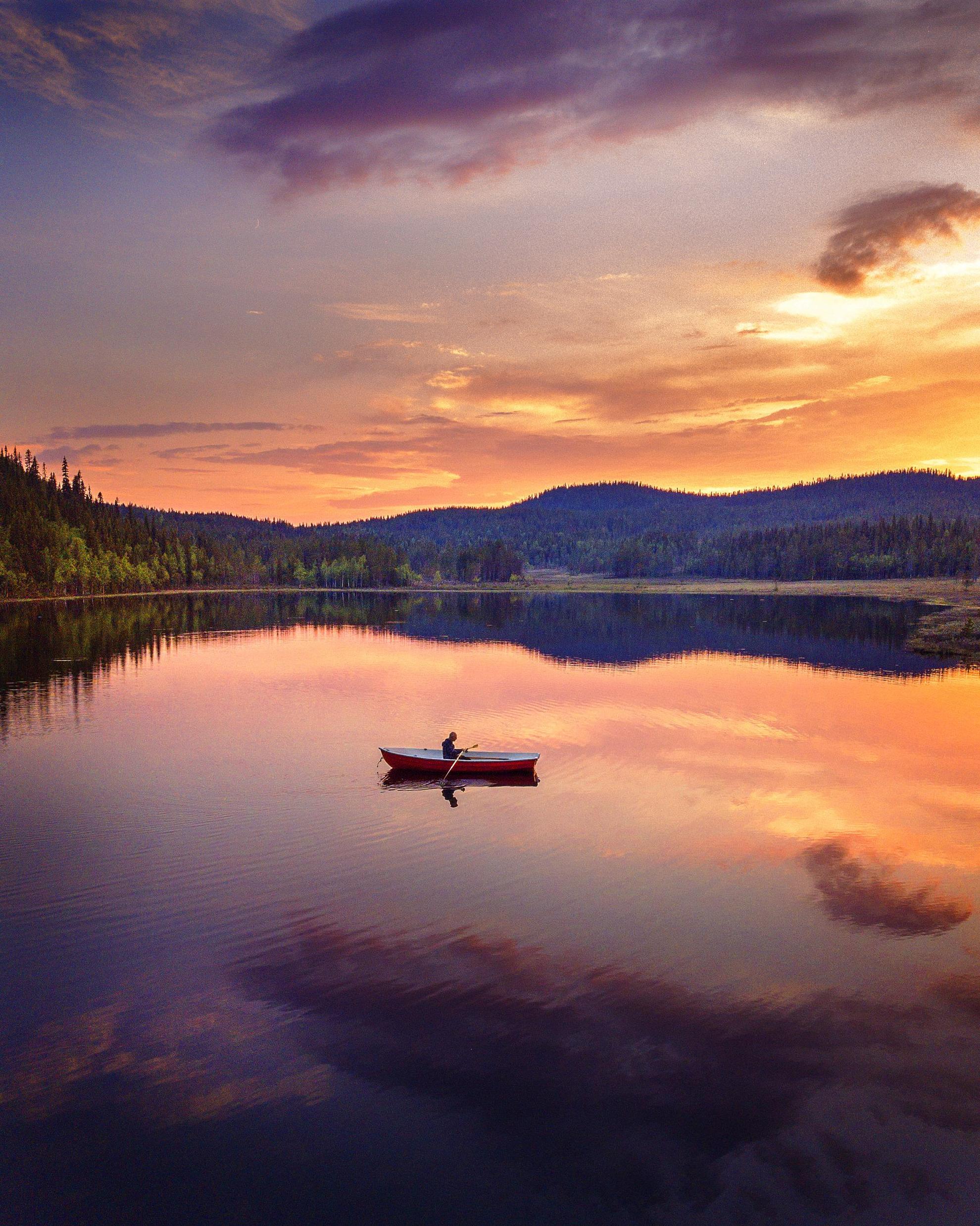 Eine Person sitzt in einem Boot und geleitet über einen See. Im Hintergrund ist ein Gebirge zu sehen und das Bild wurde in der Nacht in Schwedisch Lappland aufgenommen, weshalb die Sonne die Umgebung in orangenes Licht wirft..